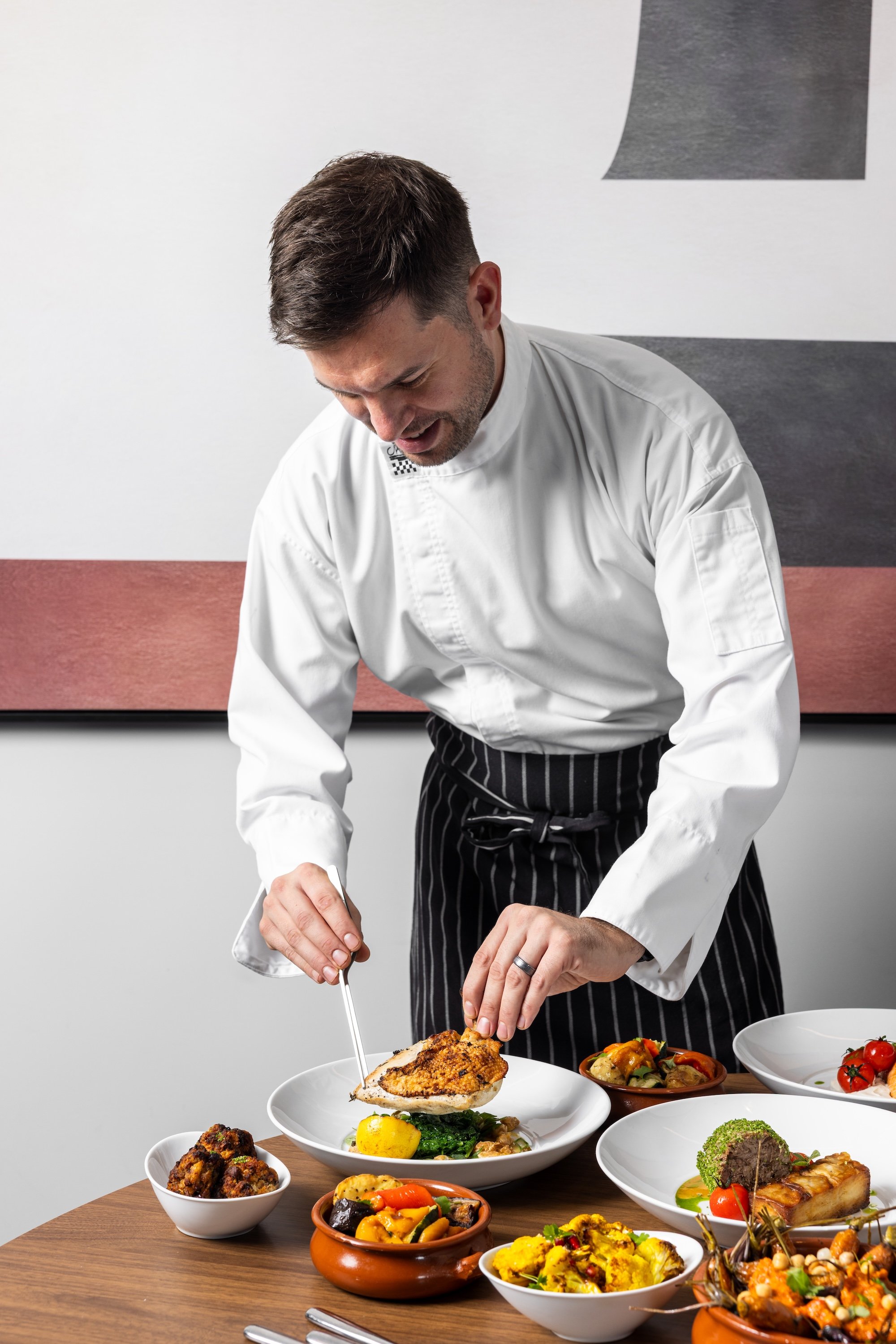 Chef garnishing a plated dish in a professional kitchen with multiple bowls of cooked food on a wooden table.