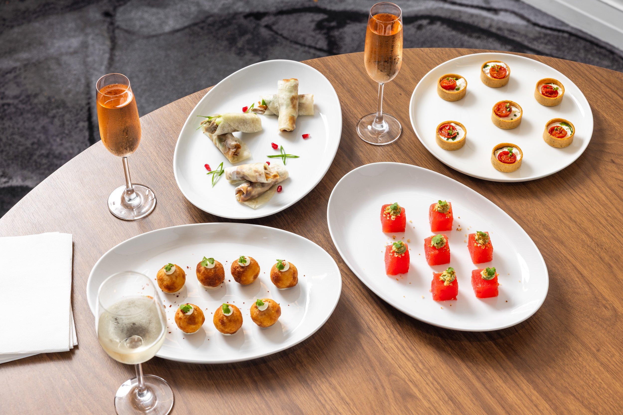 A round wooden table with four white plates of assorted appetizers and two glasses of wine, one red and one white. The appetizers include spring rolls, small topped cubes, meatballs, and watermelon cubes with garnishes.