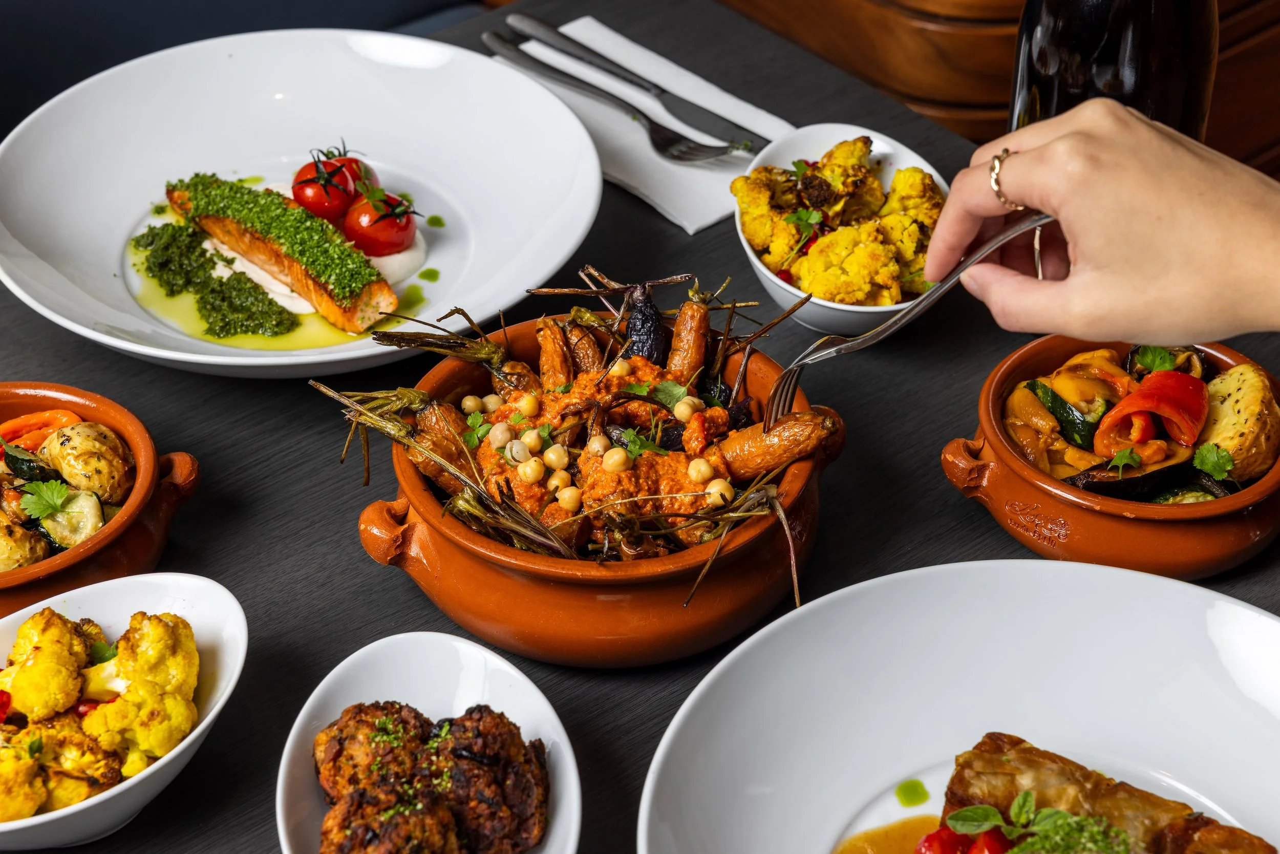 A variety of Indian dishes on a black table, including a terracotta bowl of curry with lobster, a white plate of grilled fish with cherry tomatoes, small bowls of vegetable dishes, and a person serving themselves from a bowl of cauliflower and other vegetables.