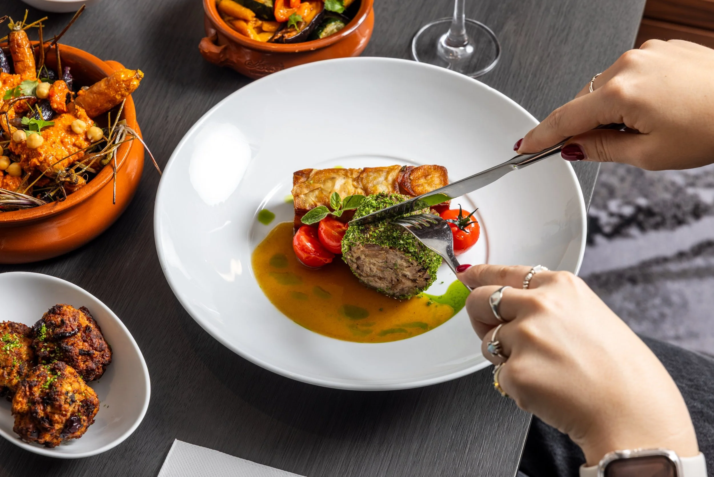 Person cutting into a meatloaf with cherry tomatoes and green sauce on a white plate, surrounded by bowls of other dishes on a dark table.