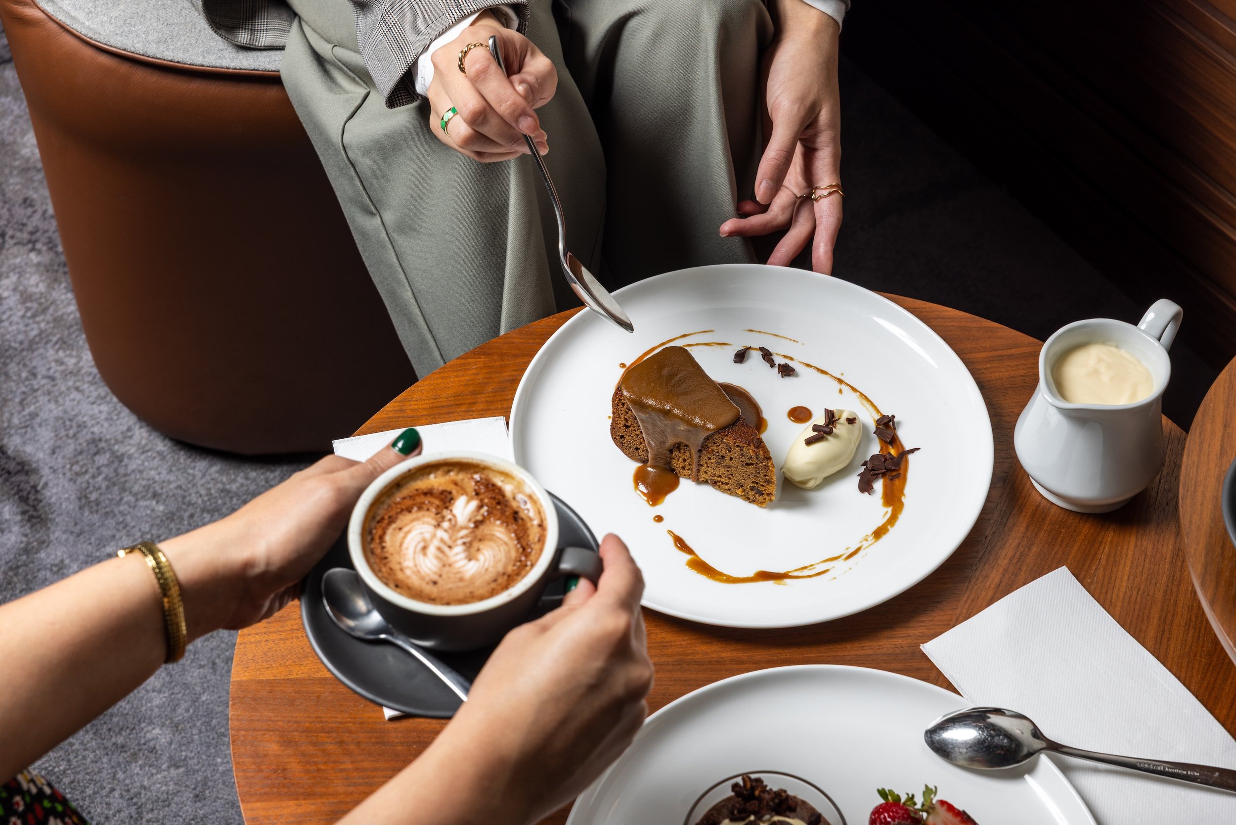 People enjoying desserts at a restaurant, including a chocolate cake with caramel sauce, a scoop of vanilla ice cream, and a cup of coffee with foam art.