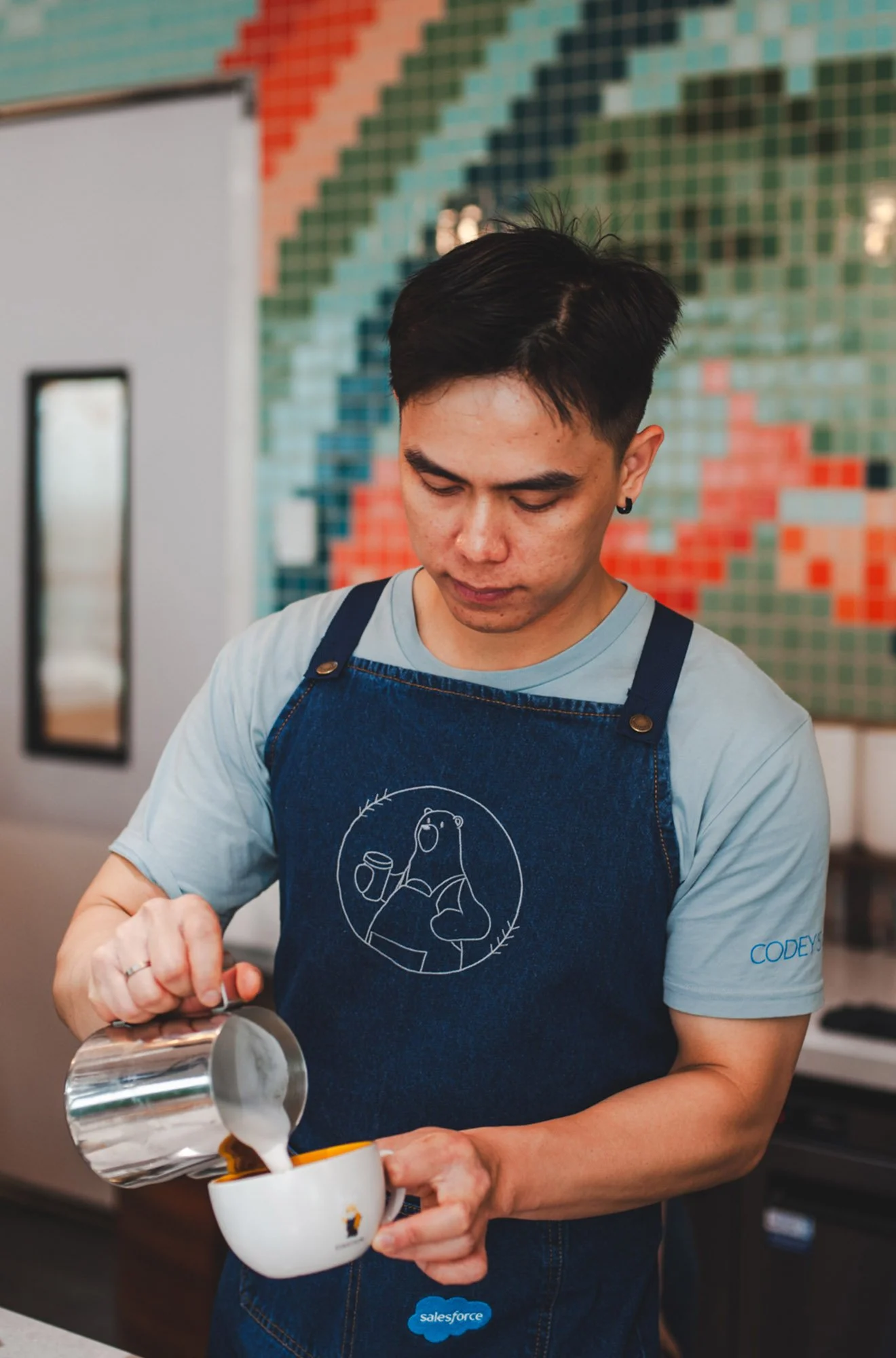 A young man with short dark hair, wearing a light blue t-shirt and a dark apron with a bear logo, pouring coffee from a stainless steel pitcher into a white mug in a colorful, modern cafe.
