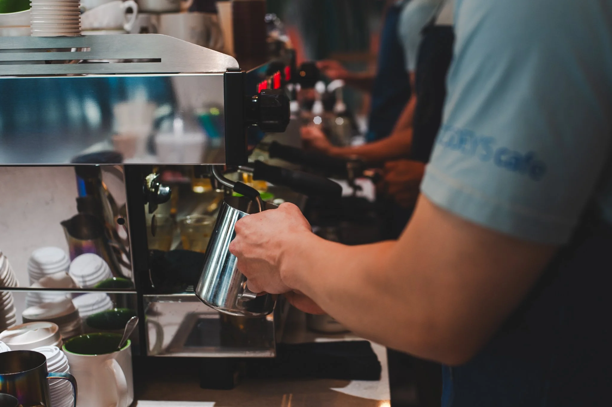A barista using a stainless steel milk frothing pitcher to steam milk at a coffee shop espresso machine, with cups, saucers, and small bowls nearby.