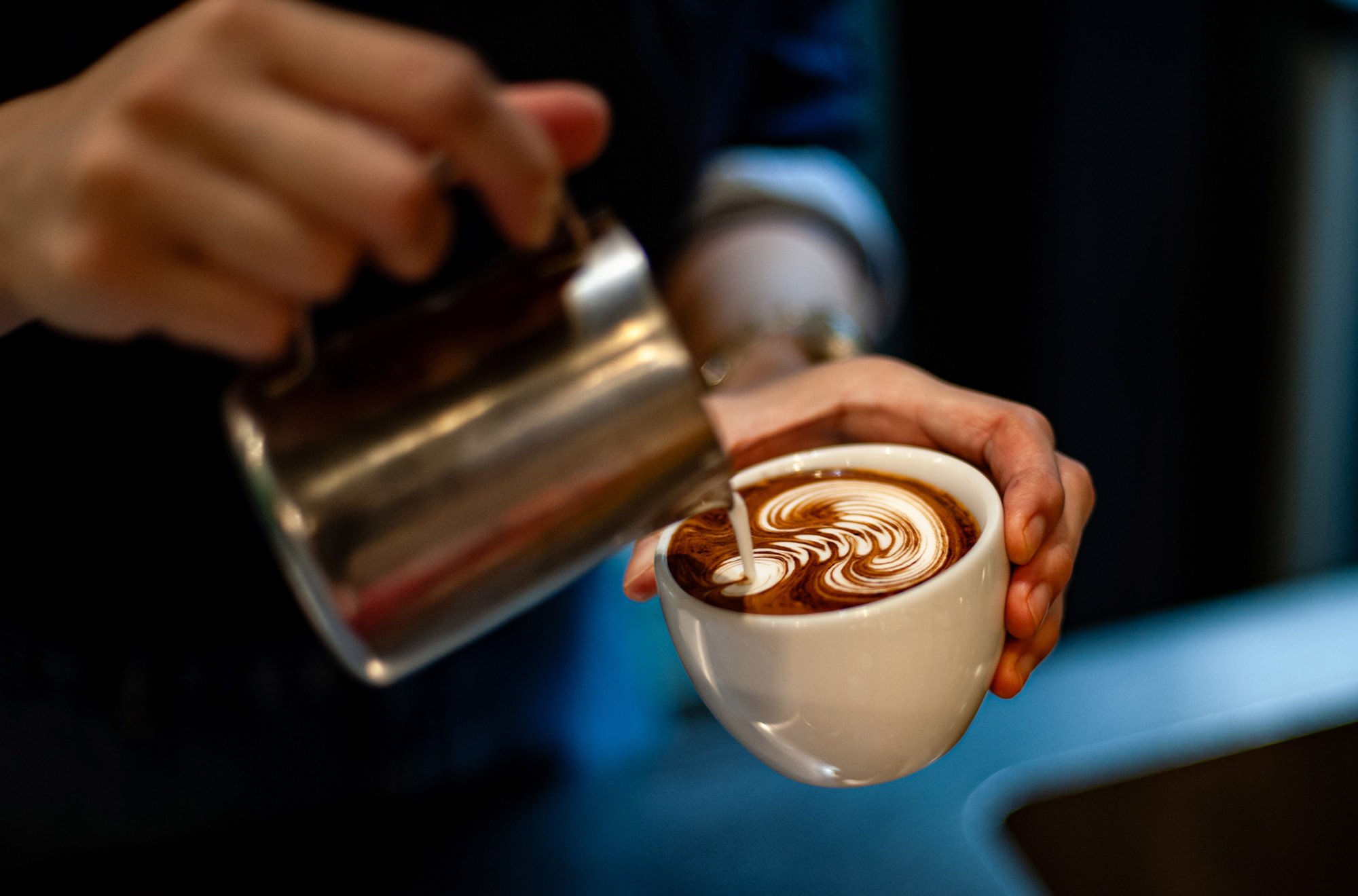 Barista pouring steamed milk into a cup of hot chocolate or coffee, creating latte art with swirling patterns.