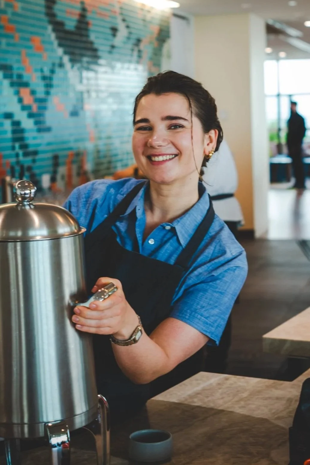 Smiling female barista with dark hair in a bun, wearing a blue shirt and black apron, standing at coffee bar, holding coffee urn, in a modern café with colorful tiled wall in the background.