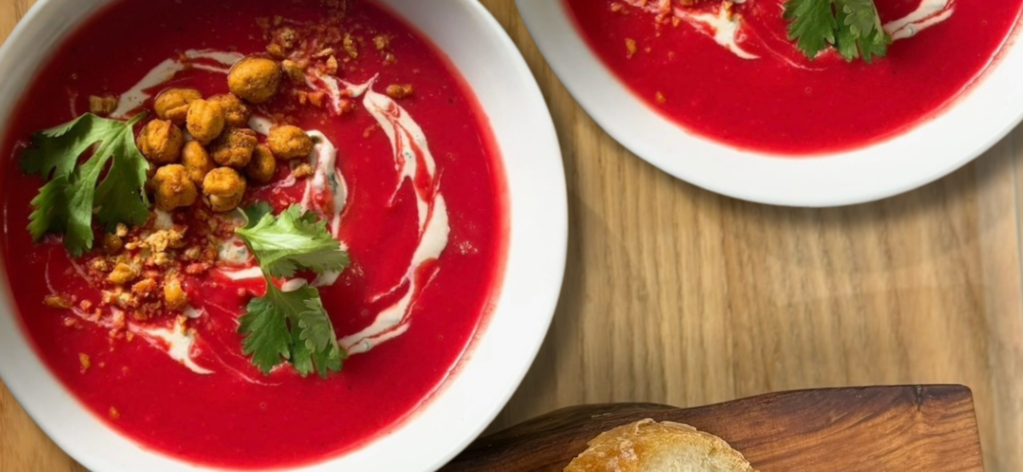 Two white bowls of vibrant red tomato soup garnished with cilantro leaves, cream swirls, and crispy chickpeas, on a wooden table with a bread roll nearby.