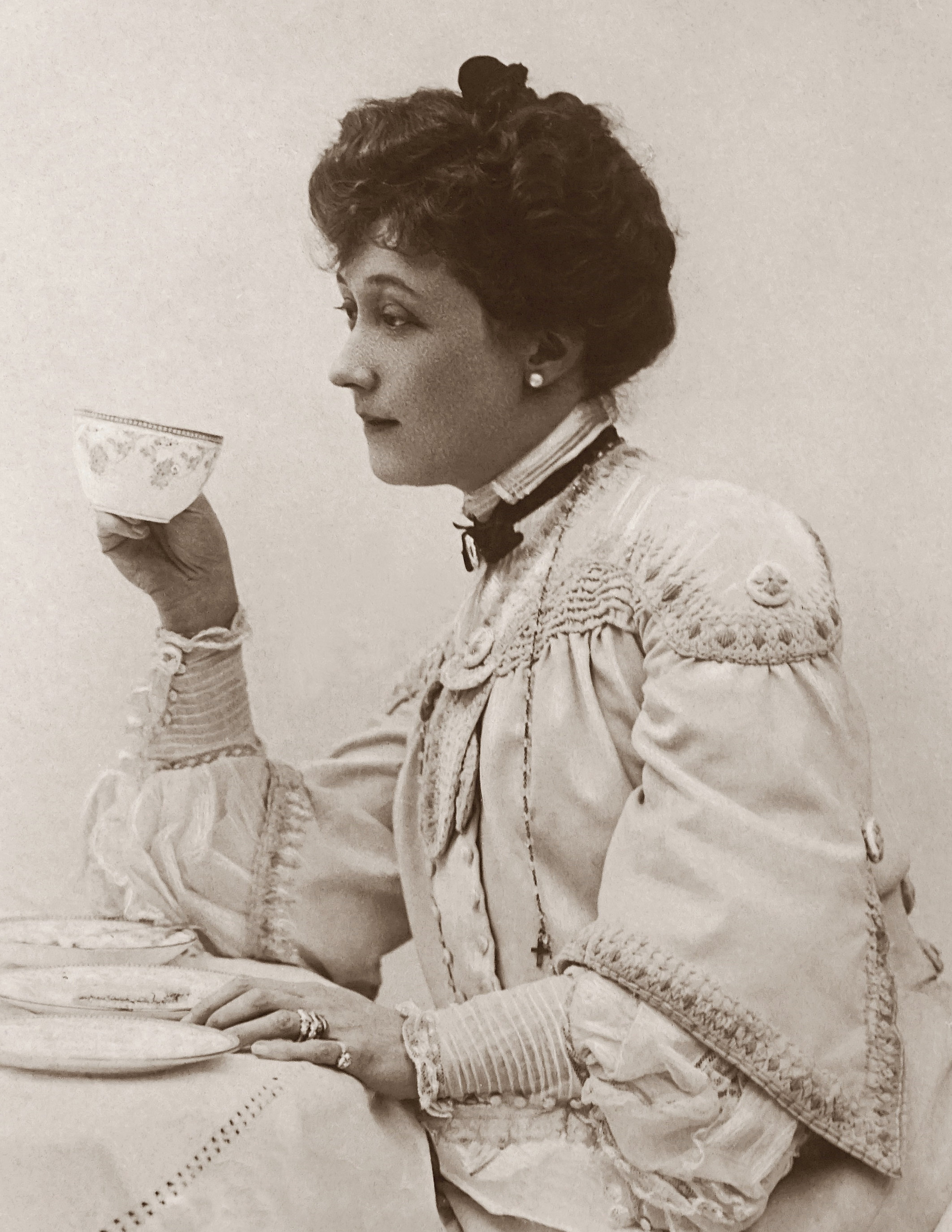 A Victorian woman holds a teacup. She wears her hair up, pearl earrings, and she is shown facing left.