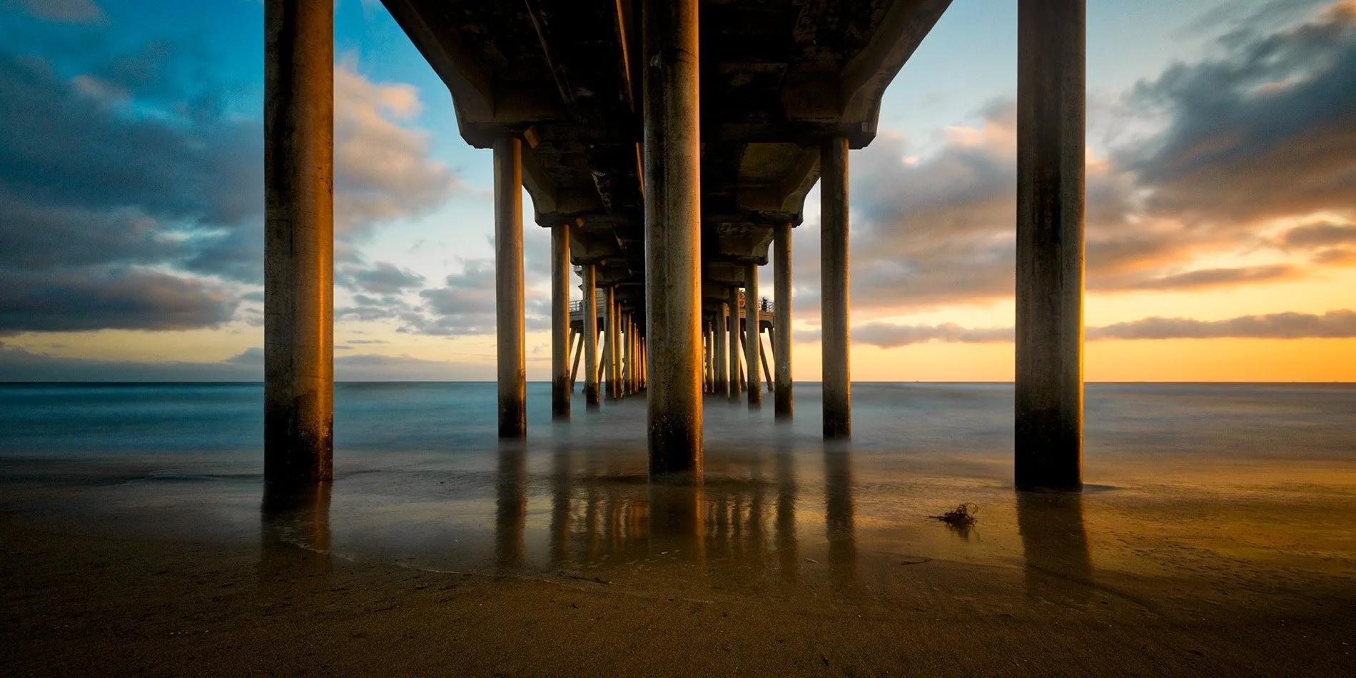 Underneath a pier with concrete columns at sunset, water and sand visible.