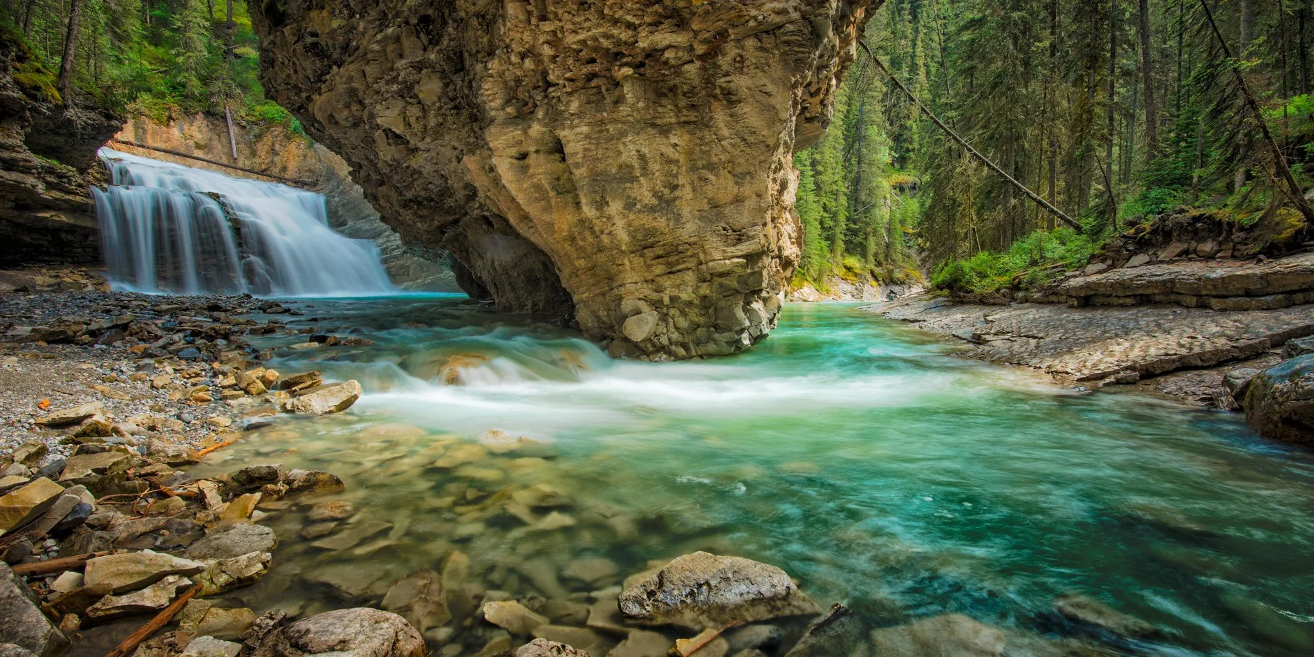 A flowing river with a small waterfall, surrounded by rocks and dense green forest, with a large rock formation overhanging the water.