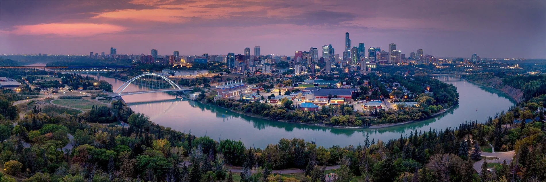 A city skyline at sunset with a river winding through a park in the foreground. The city is illuminated with tall buildings and a bridge over the river.