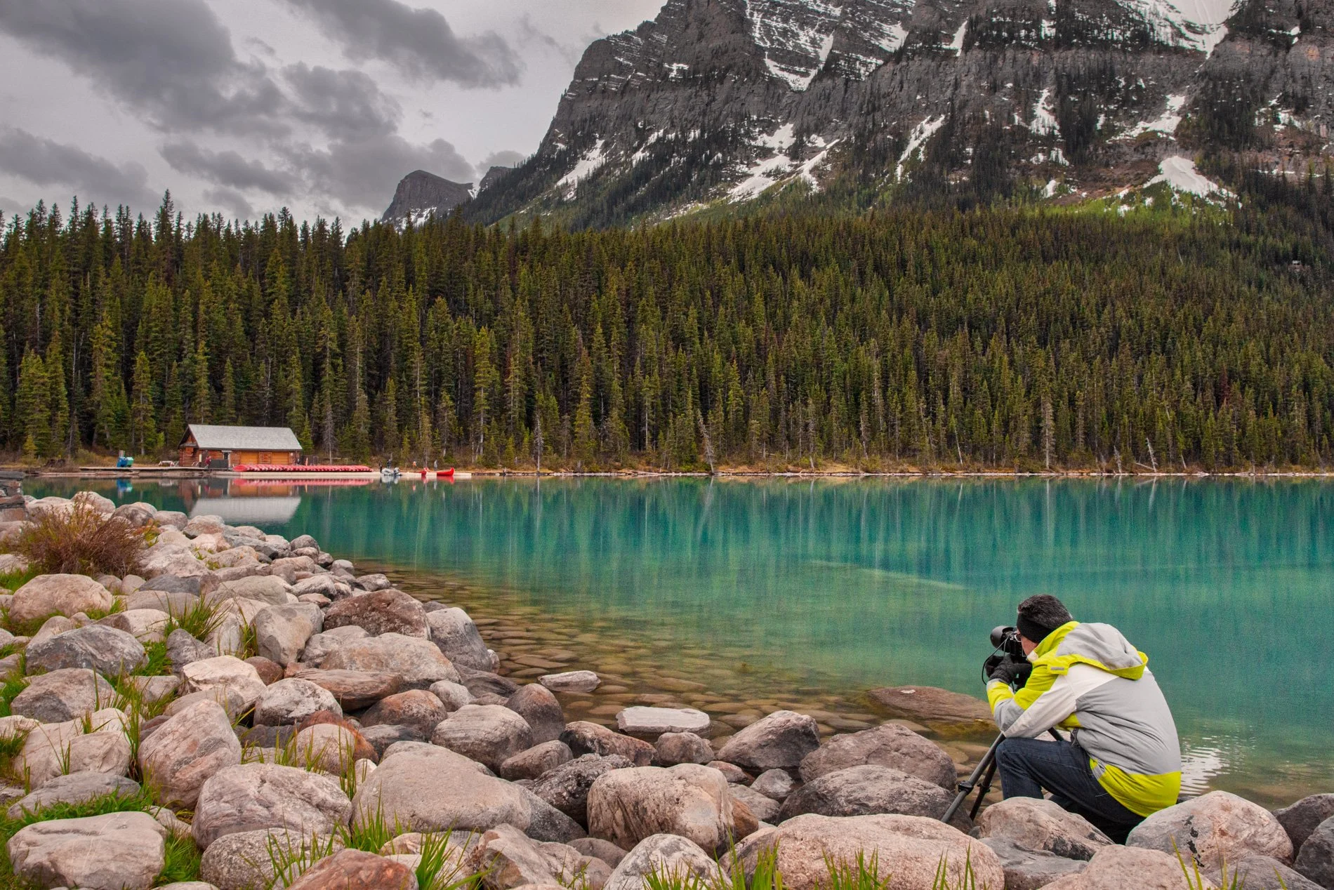 A person wearing a gray and yellow jacket is using a camera on a tripod at the rocky shore of a greenish-blue lake, with a dense forest, mountains with snow patches, and cloudy sky in the background.