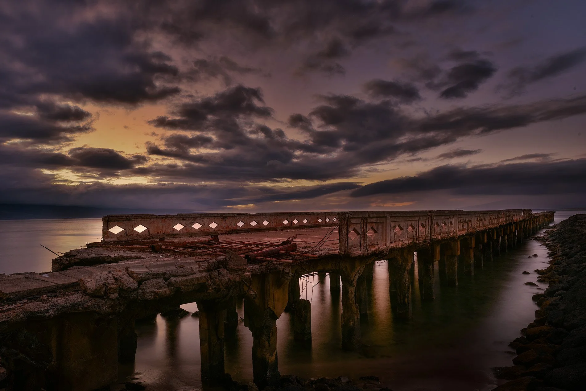 A dilapidated pier extends into the ocean under a moody, cloudy sunset sky.