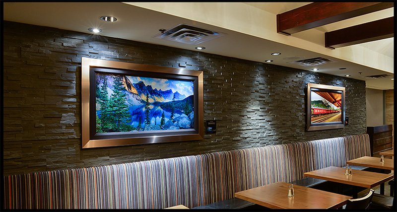 Interior of a restaurant with a stone wall, two framed landscape pictures, a striped booth, and wooden tables with small vases.