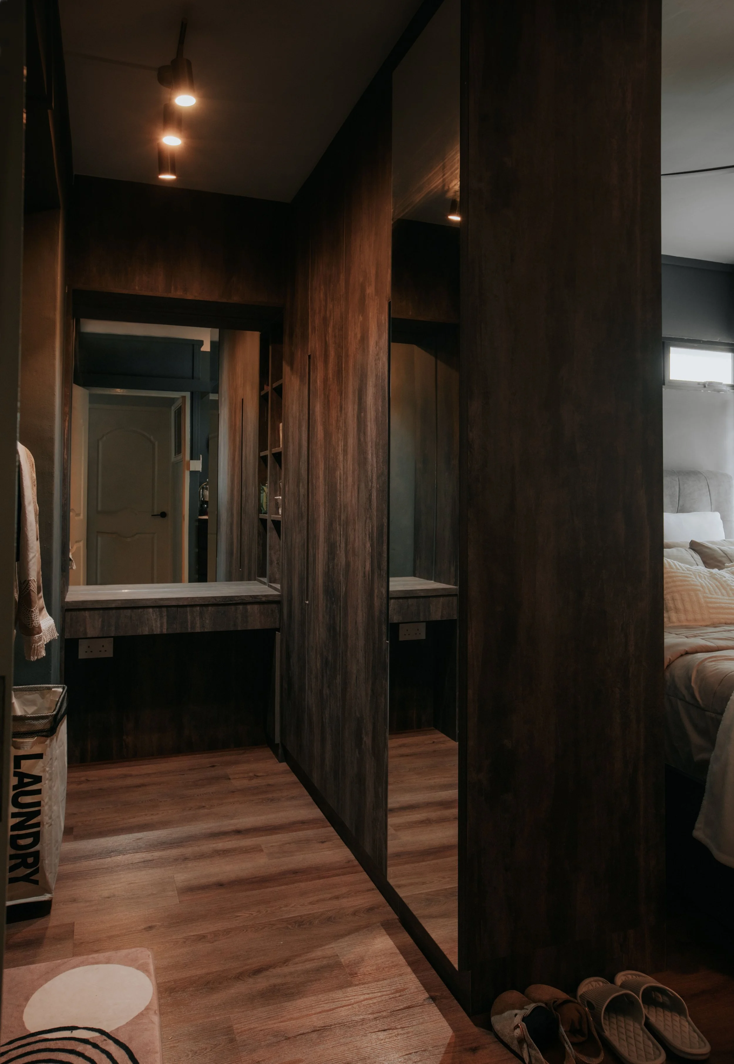 A dark wooden partition separates a bedroom from a vanity area with mirrors and power outlets. The bedroom has a bed with pillows and a window, and there are slippers on a wooden floor.