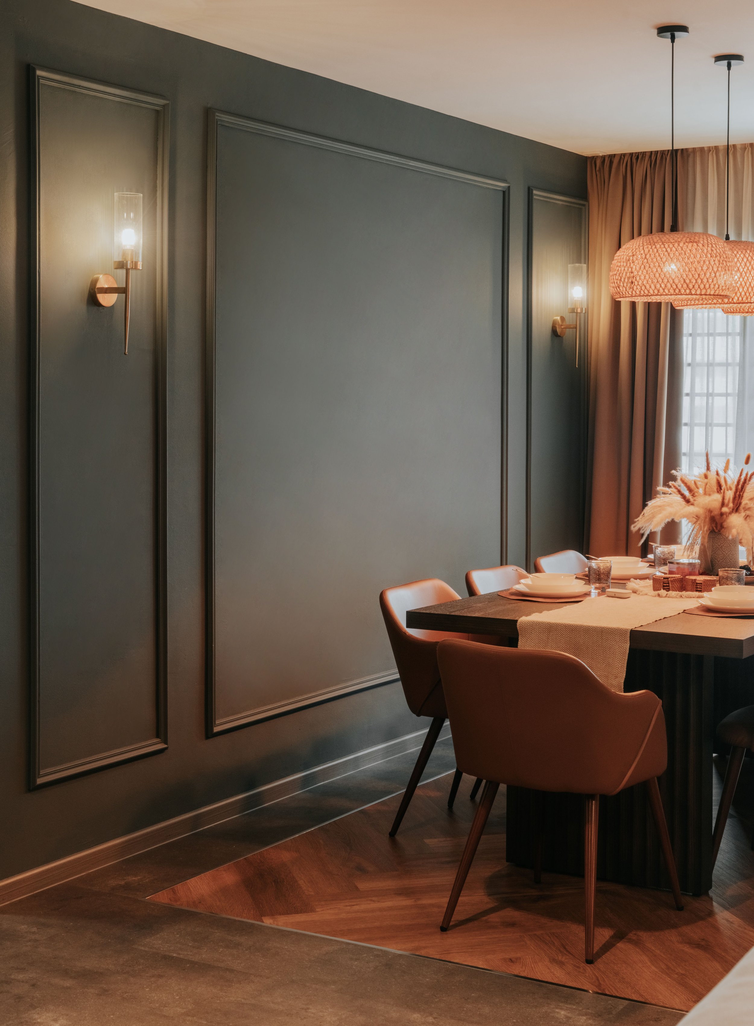Elegant dining room with a dark wooden table, pink chairs, tableware, floral centerpiece, wall sconces, pendant light, and window with beige curtains.