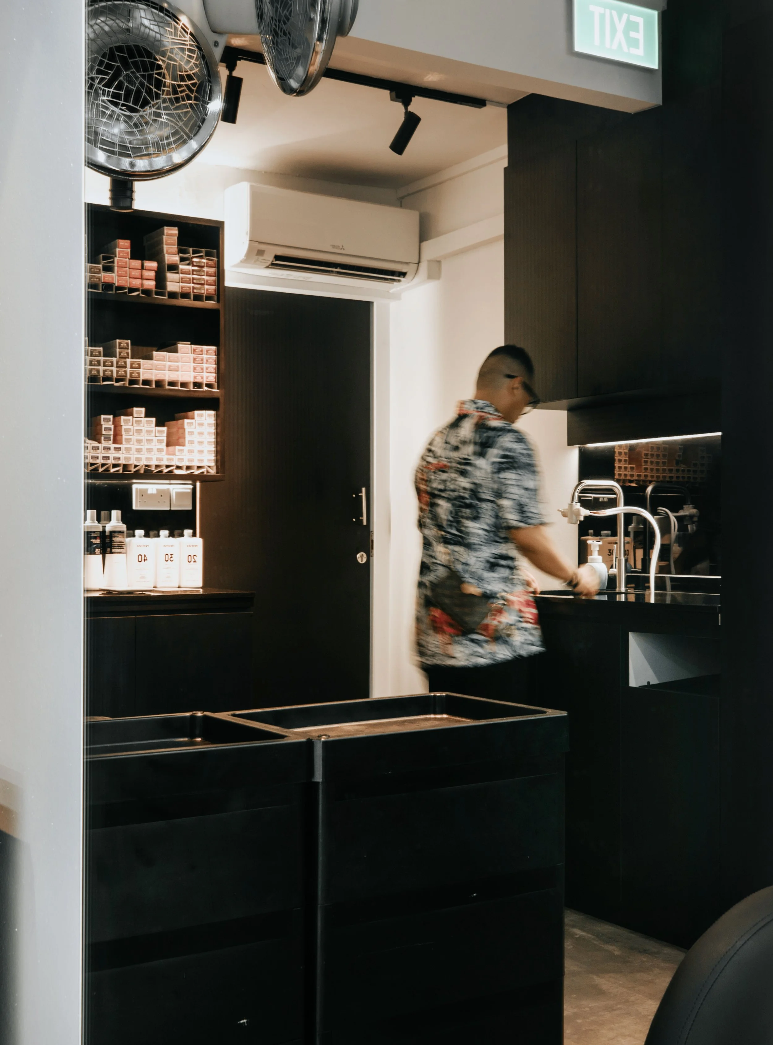 A man wearing a patterned shirt is washing dishes at a black sink in a modern kitchen. The background includes shelves with spice jars, bottles, and a black door. The ceiling has a mounted air conditioner, a fan, and a spotlight. An illuminated exit 