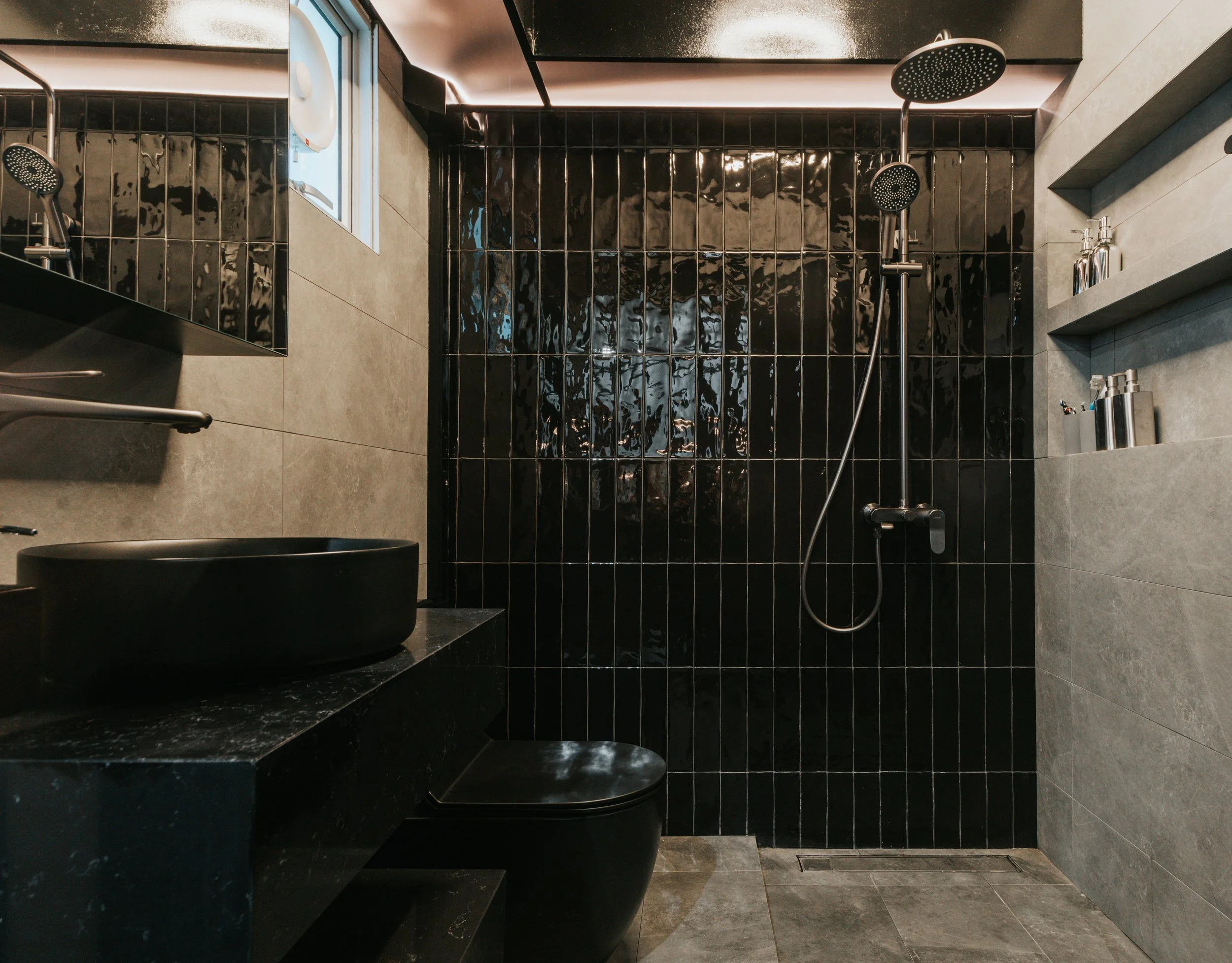 Modern bathroom with black and gray tiles, a black vessel sink on a black marble countertop, a black toilet, and black shower fixtures on a black tiled wall, with gray tiled walls and shelves holding bottles.