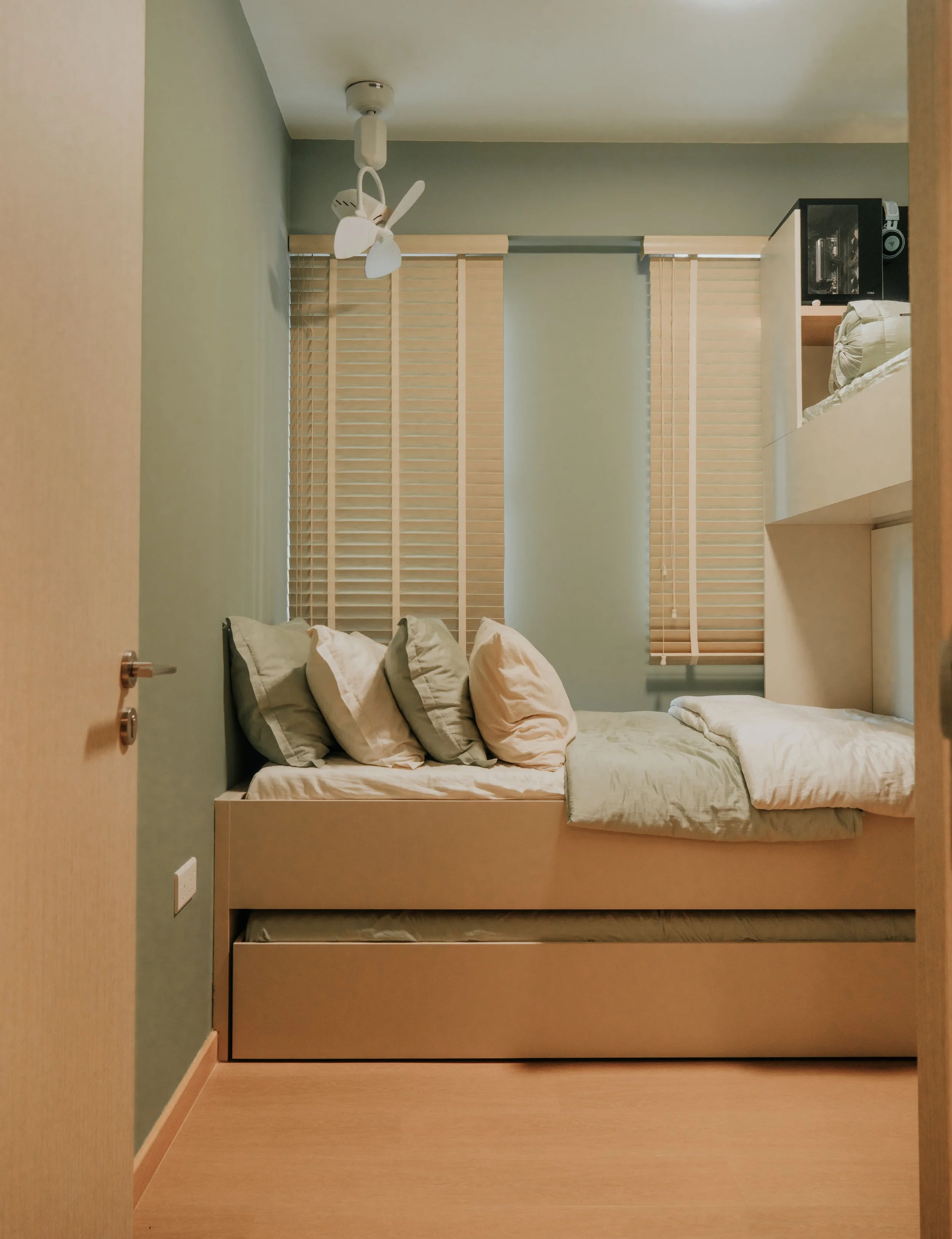 A small bedroom with a bed that has multiple pillows, beige walls, and wooden blinds on the window. There is a ceiling fan with lights, and a black microwave on a shelf in the corner.