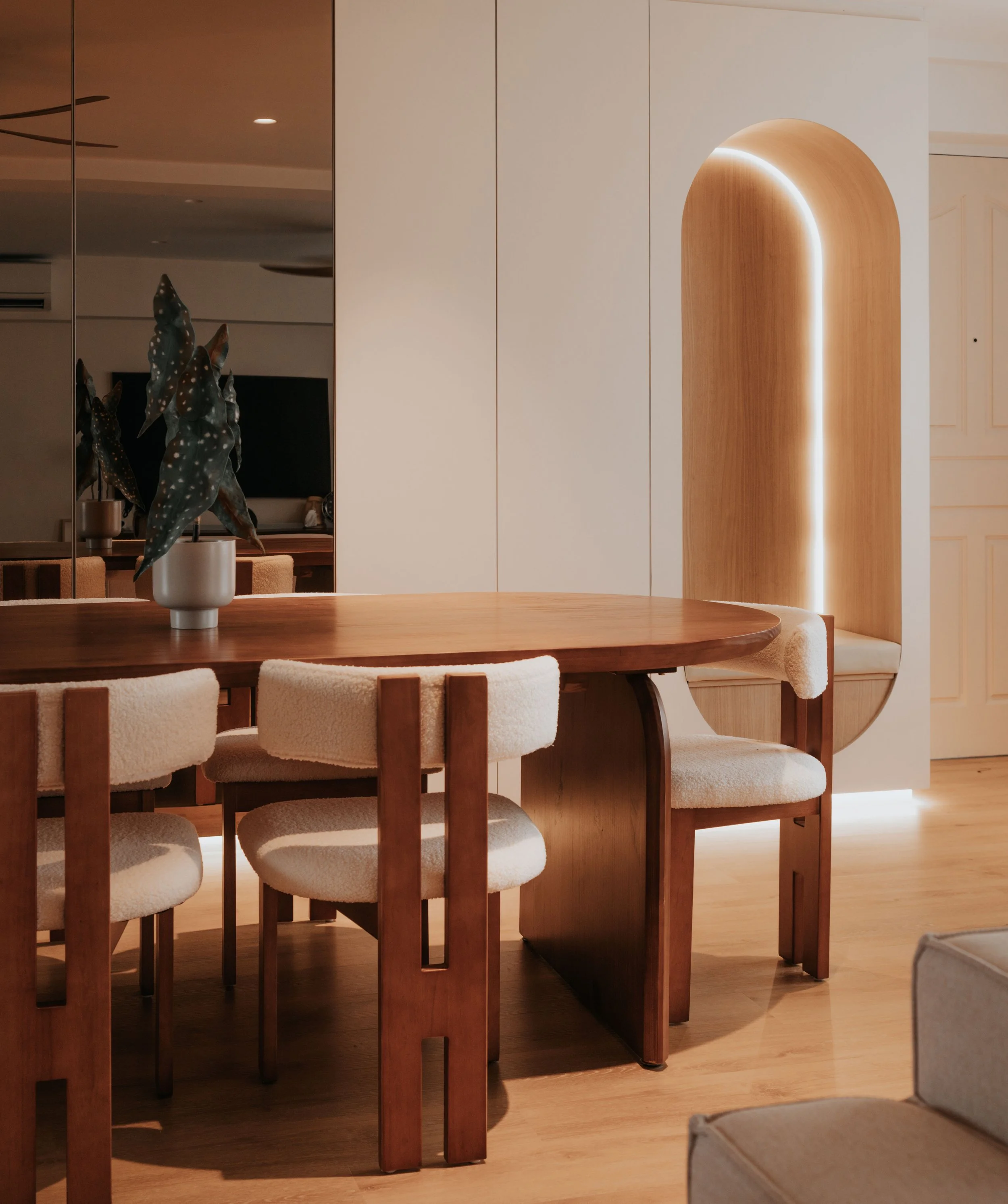 A wooden dining table with four upholstered chairs and a potted plant in a modern room with warm lighting and minimalist decor.