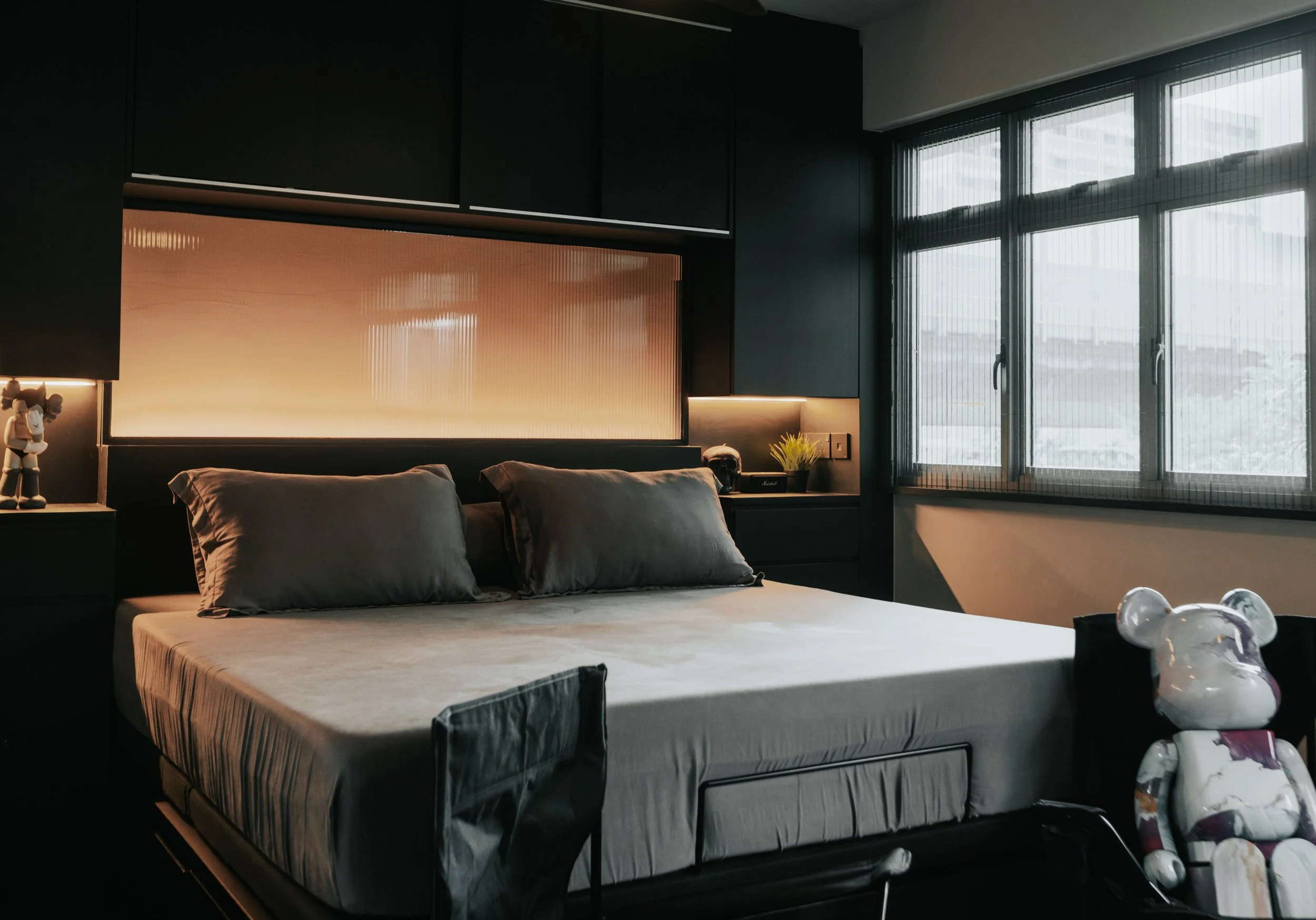 Modern bedroom with dark cabinetry, a large bed with grey pillows, and a side window with sheer curtains. There are decorative items, including a toy bear and a small potted plant, near the window.