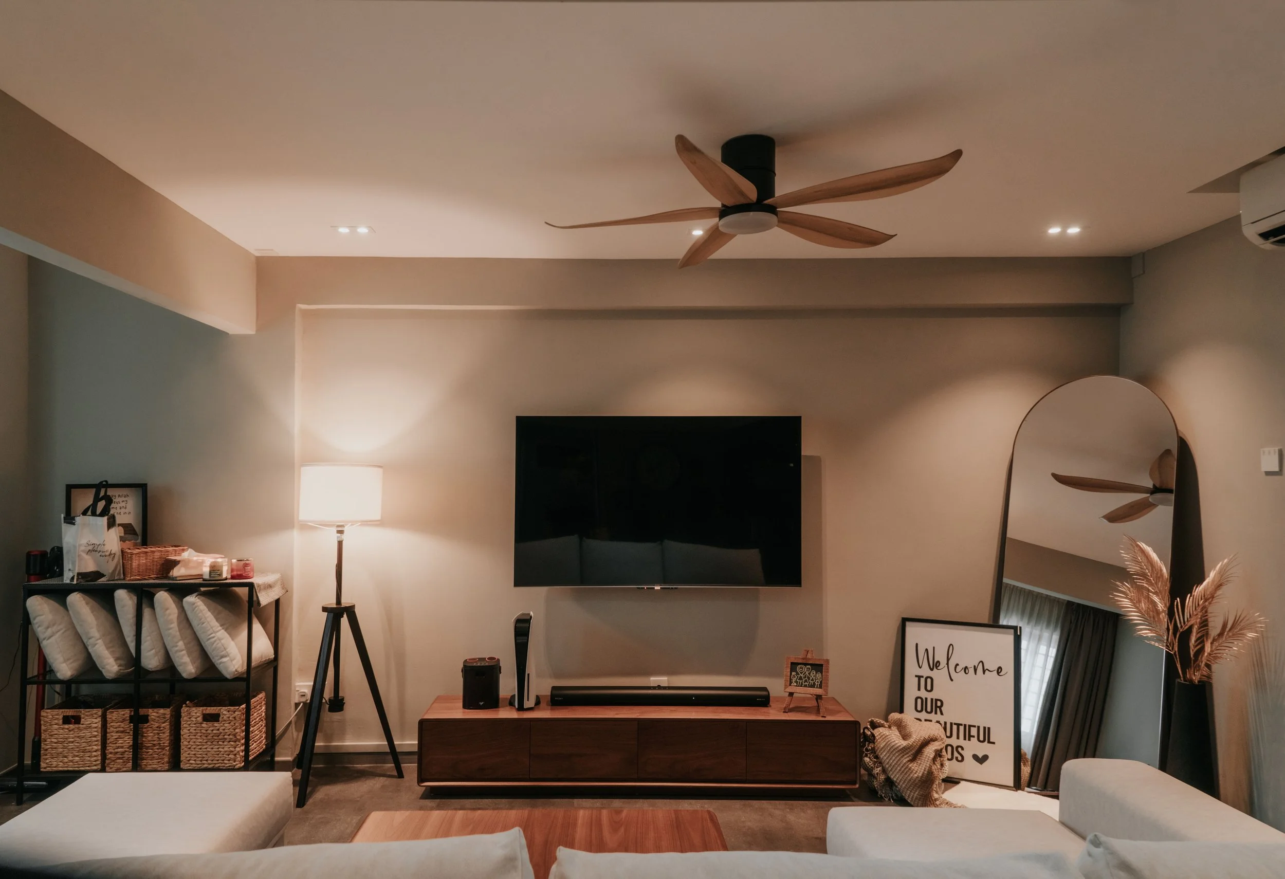 Living room with a flat-screen TV mounted on the wall, a wooden media console below, a standing lamp on the left, a large mirror leaning against the right wall, a decorative sign next to the mirror, a ceiling fan with wooden blades, and various decor
