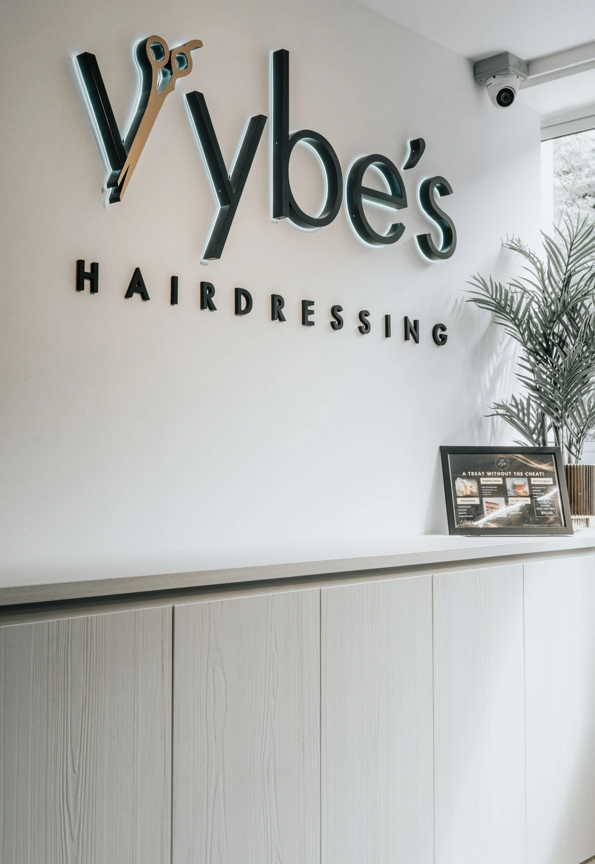 Wall sign with the words 'Y' by Y's Hairdressing, a salon with a minimalist decor, with a potted plant and a framed menu on a light-colored counter.