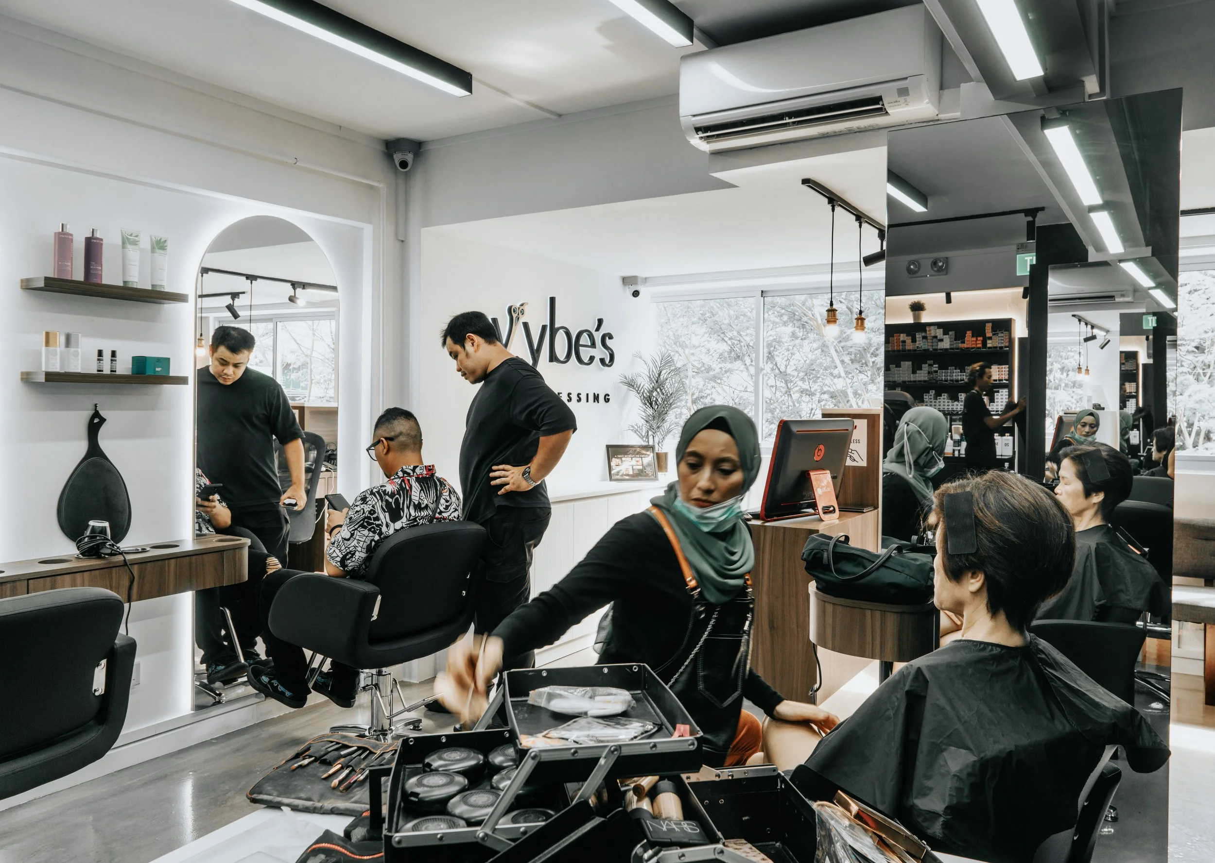 People getting haircuts and hair styling services at a modern hair salon with white walls, black furniture, and large windows with natural light.