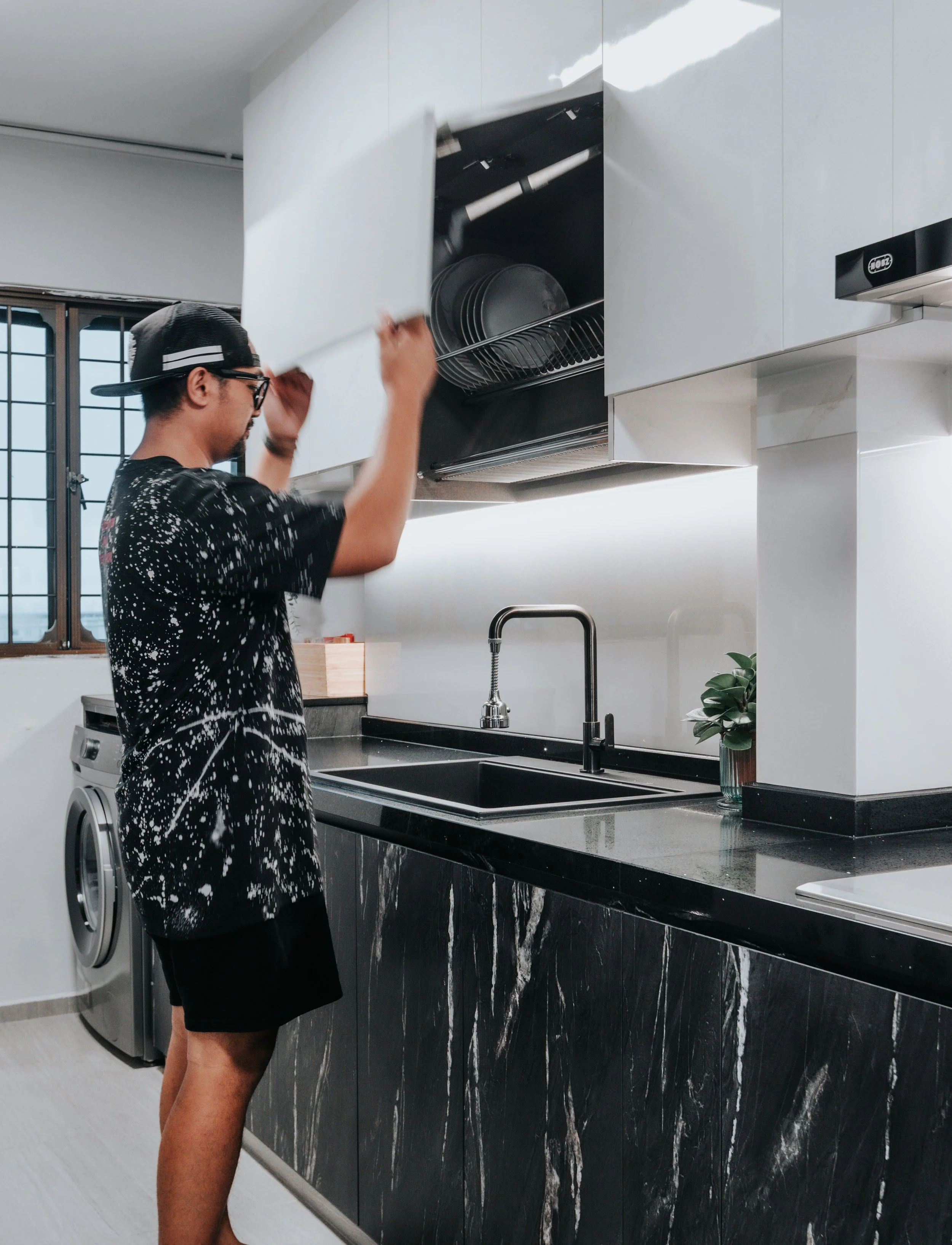 Man in a black shirt opening a cabinet in a modern kitchen with marble countertops.