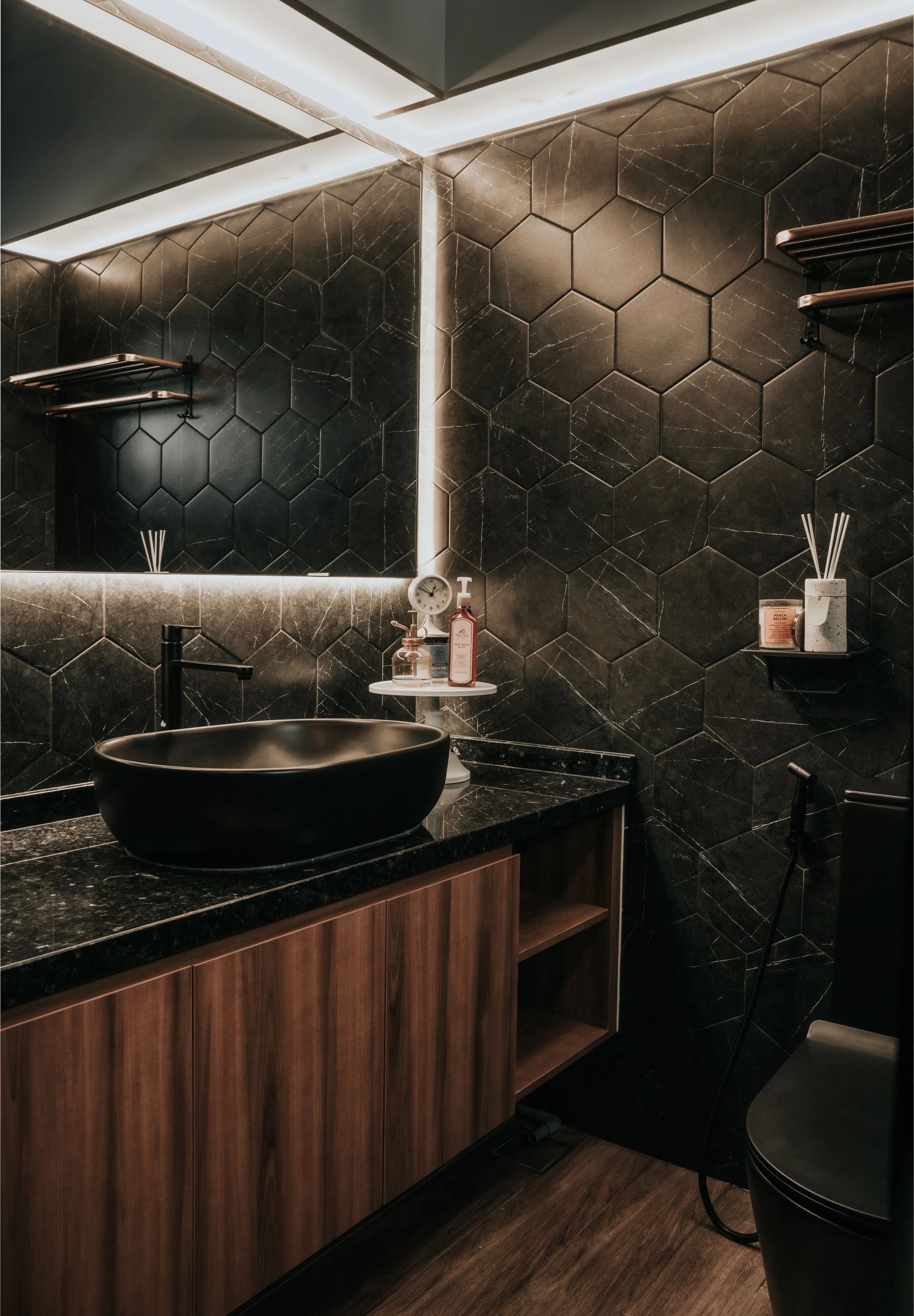 Modern bathroom with black hexagonal tiled walls, a black vessel sink on a dark granite countertop, and wooden cabinetry. Illuminated mirror, shelves with decor, and a dark toilet with a bidet spray.