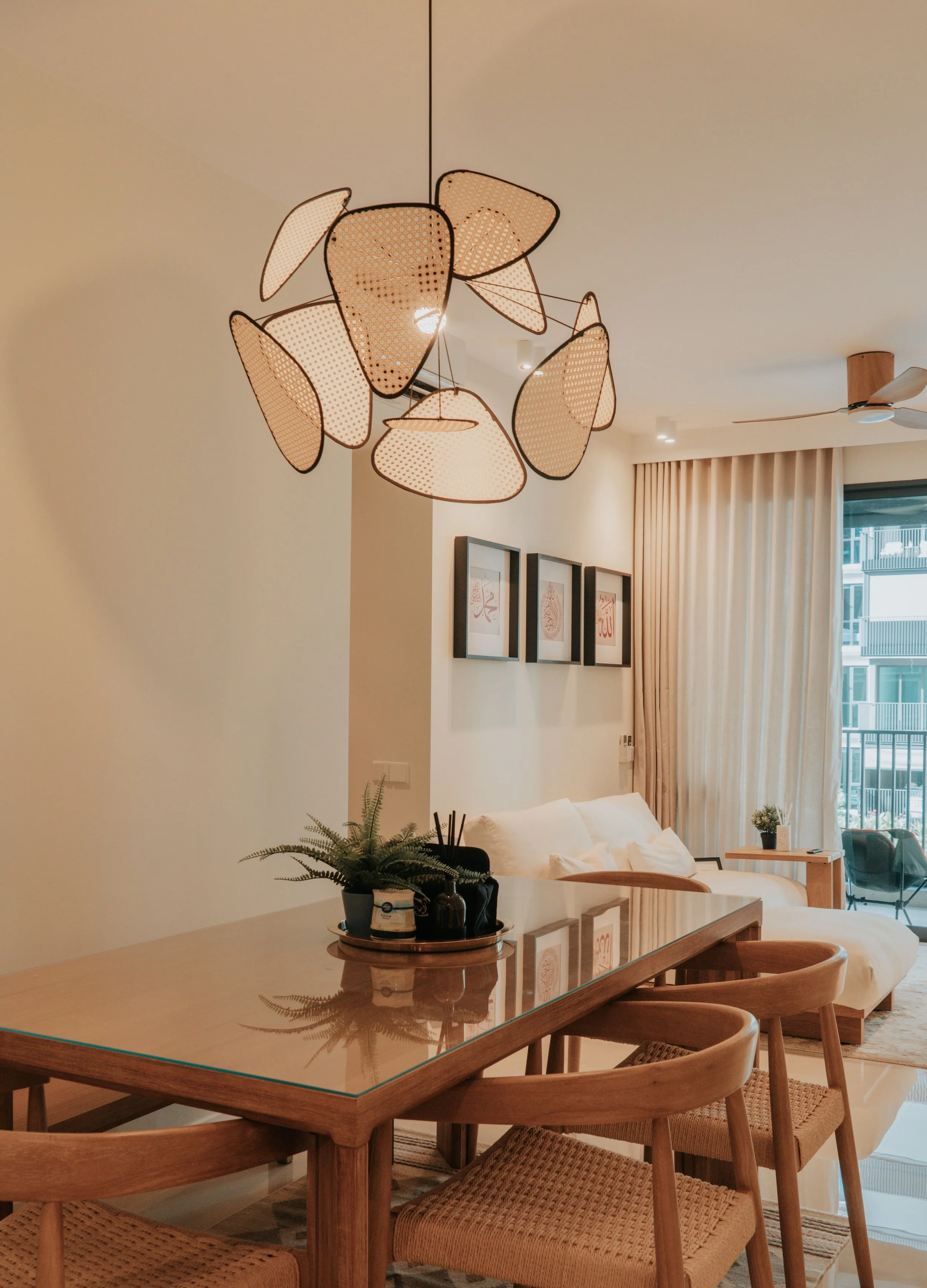 Living room with a wooden dining table, rattan chairs, a white sofa, framed artwork on the wall, potted plants, and large windows with beige curtains, illuminated by a modern hanging light fixture.