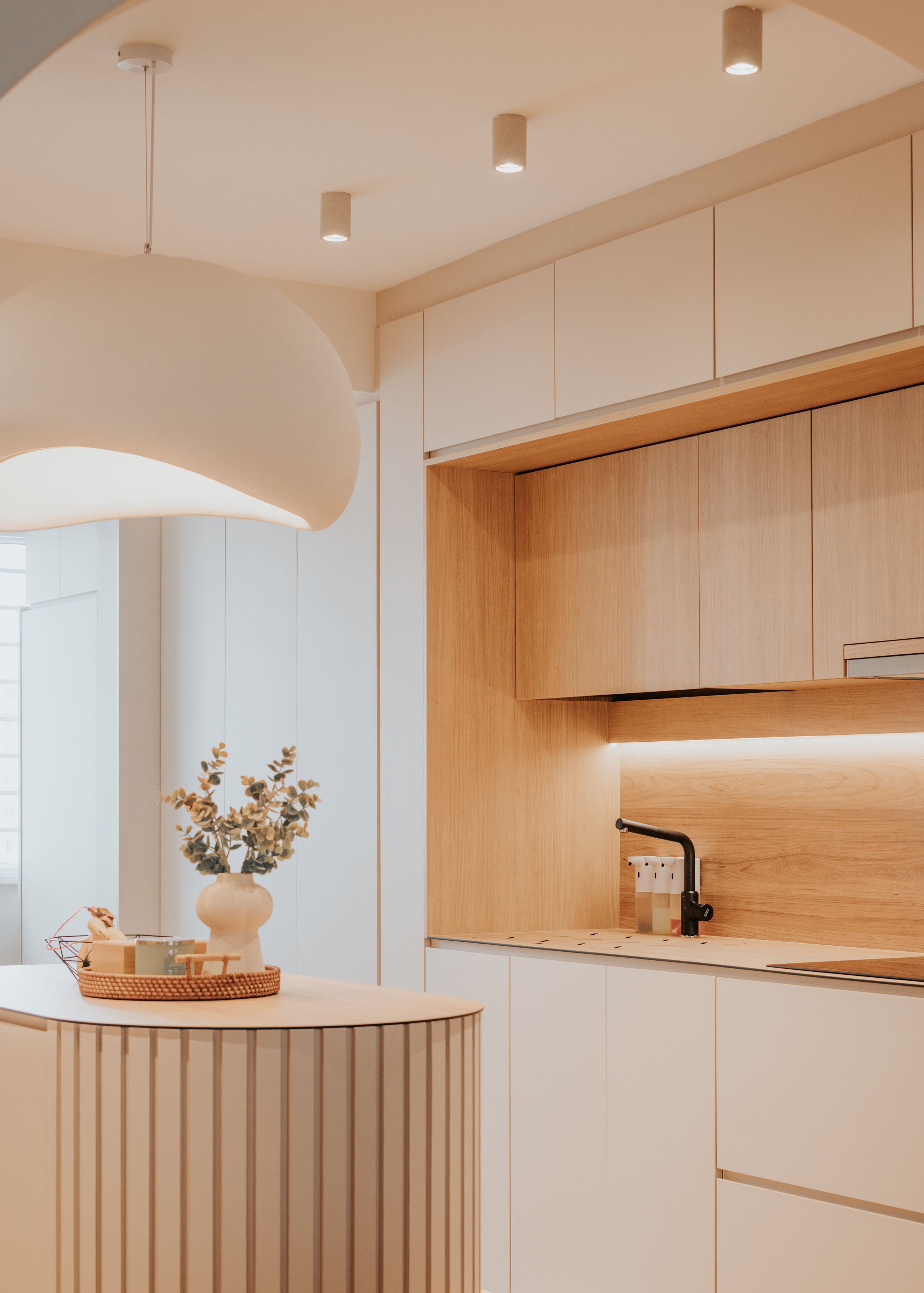 Modern kitchen with white and wood cabinetry, black faucet, and a large white pendant light, with decorative vase and tray on a curved island.