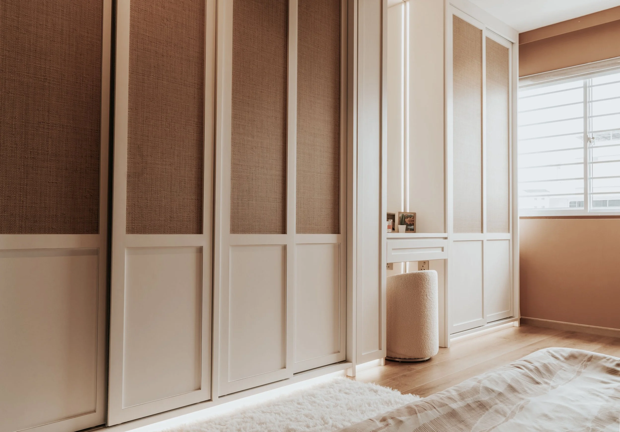 A bedroom with white built-in wardrobes with textured panels, a soft beige ottoman, a window with horizontal blinds, a fluffy white rug, and part of a bed with beige bedding.