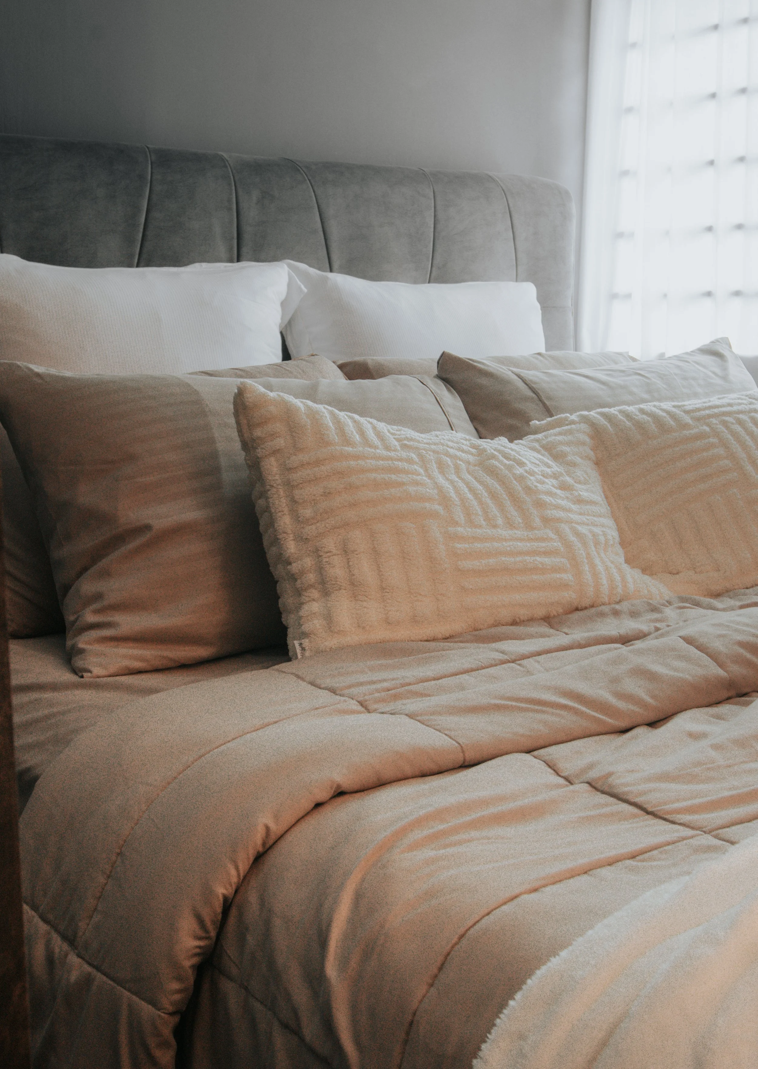 A neatly made bed with beige and white pillows and a beige quilt, in a well-lit room with a window.