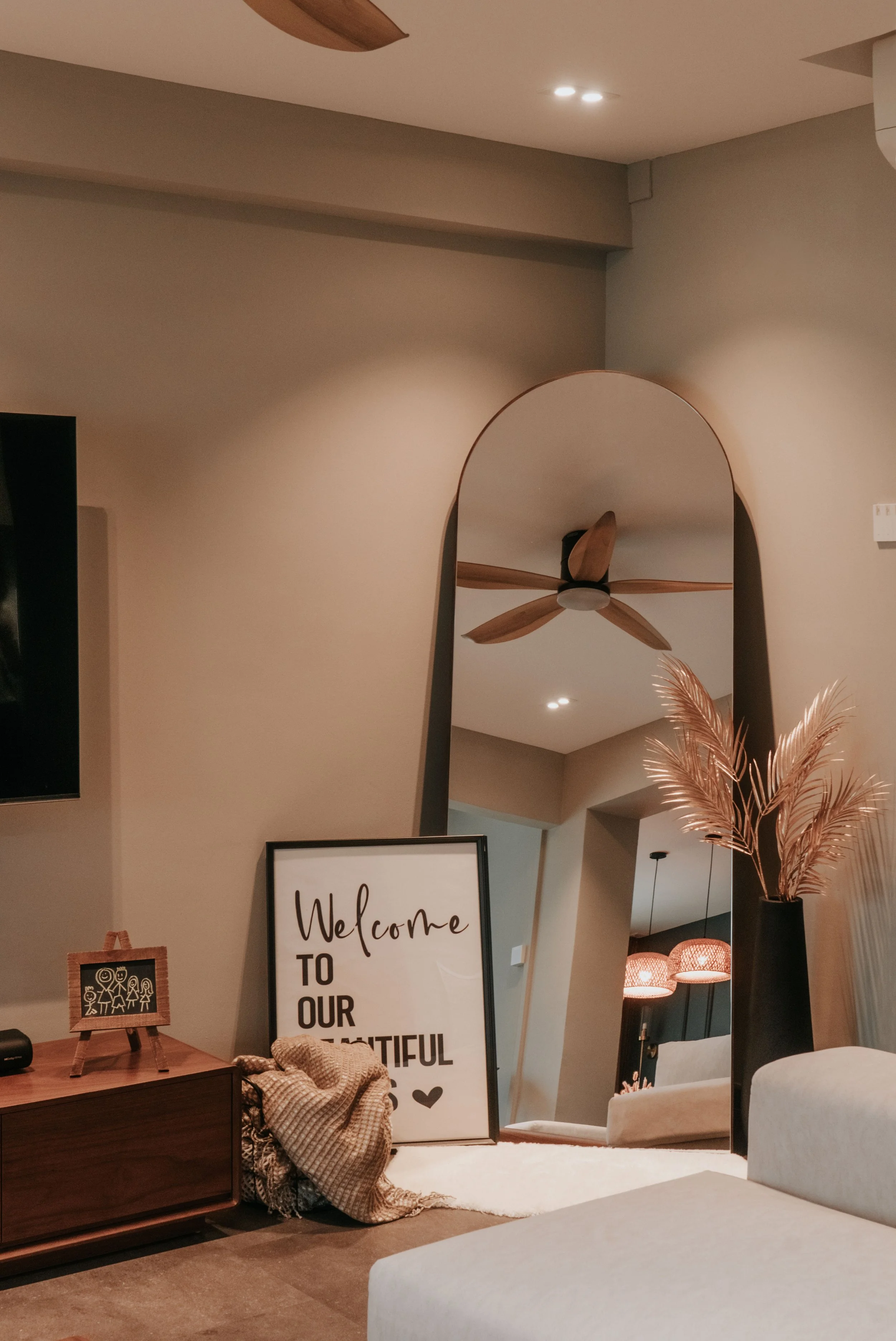 A living room corner with a large arched mirror, a wooden furniture piece, a welcome sign, a basket with a blanket, and decorative dried plants in a black vase. Part of a couch and ceiling fan with wooden blades are also visible.
