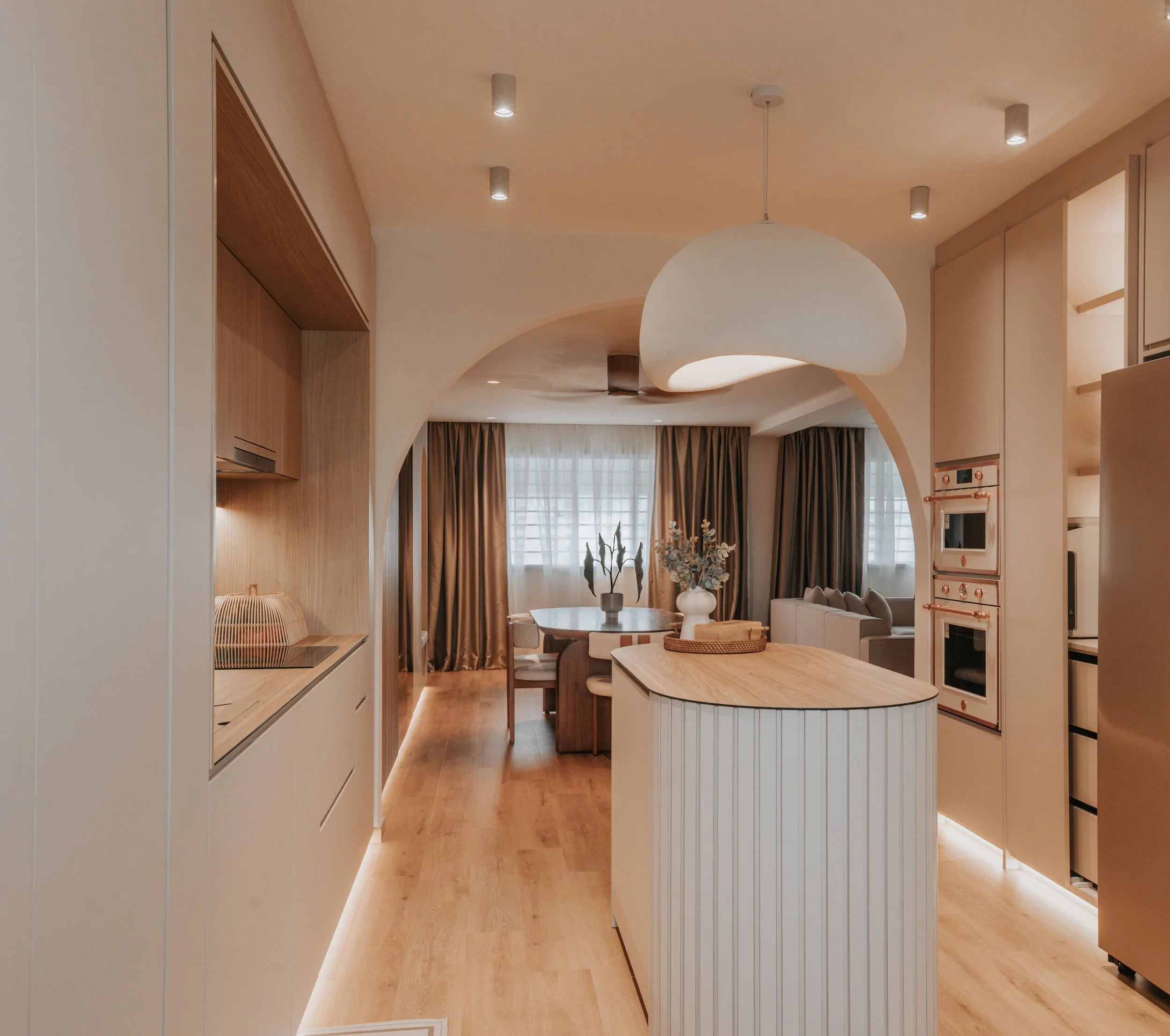 Modern open-concept kitchen and dining area with wooden flooring and neutral-toned decor, featuring a large white pendant light and a dining table with chairs near windows with curtains.