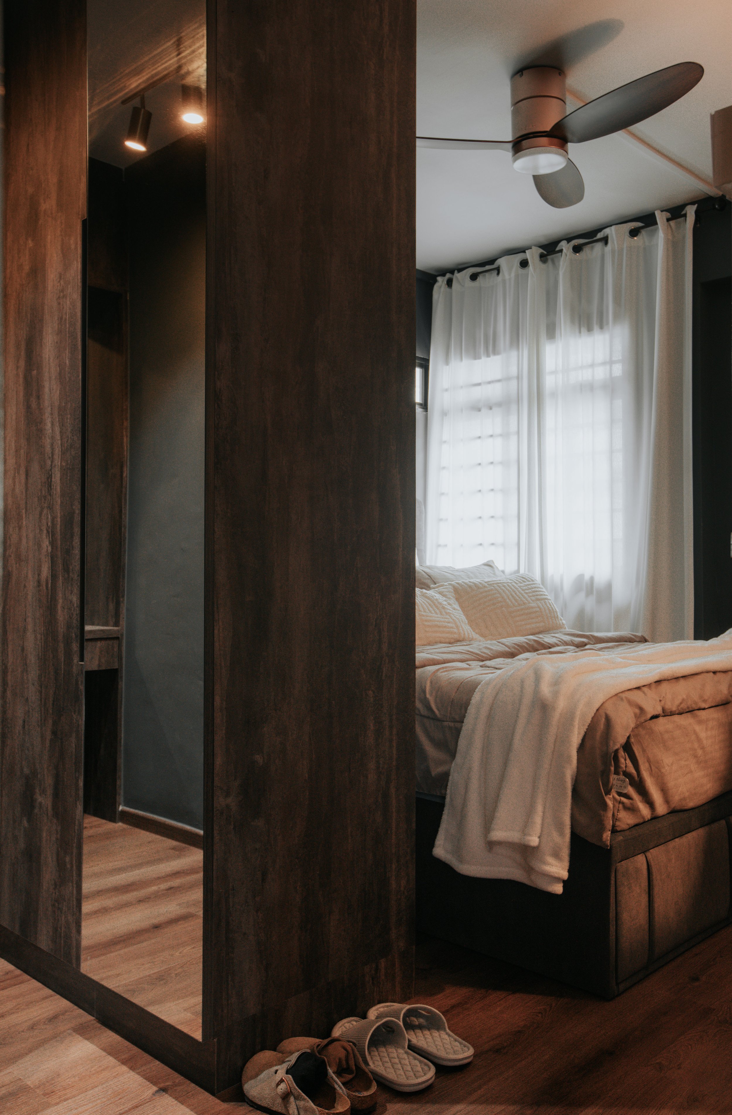 Interior of a bedroom with a dark wooden partition, a bed with beige bedding, white curtains, a ceiling fan, and slippers on the wooden floor.