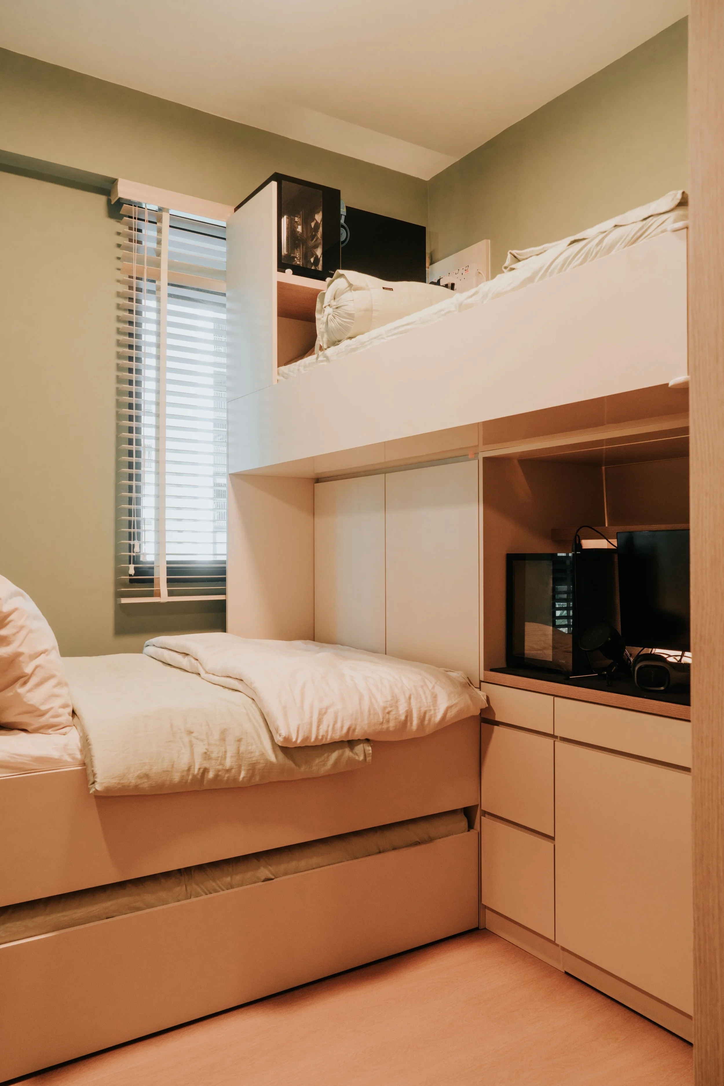 A small bedroom with a bunk bed and a window with blinds. The lower bunk has a beige mattress with a beige comforter and pillow. The upper bunk has a rolled-up blanket. There is a built-in storage unit with a TV, a microwave, and other items.