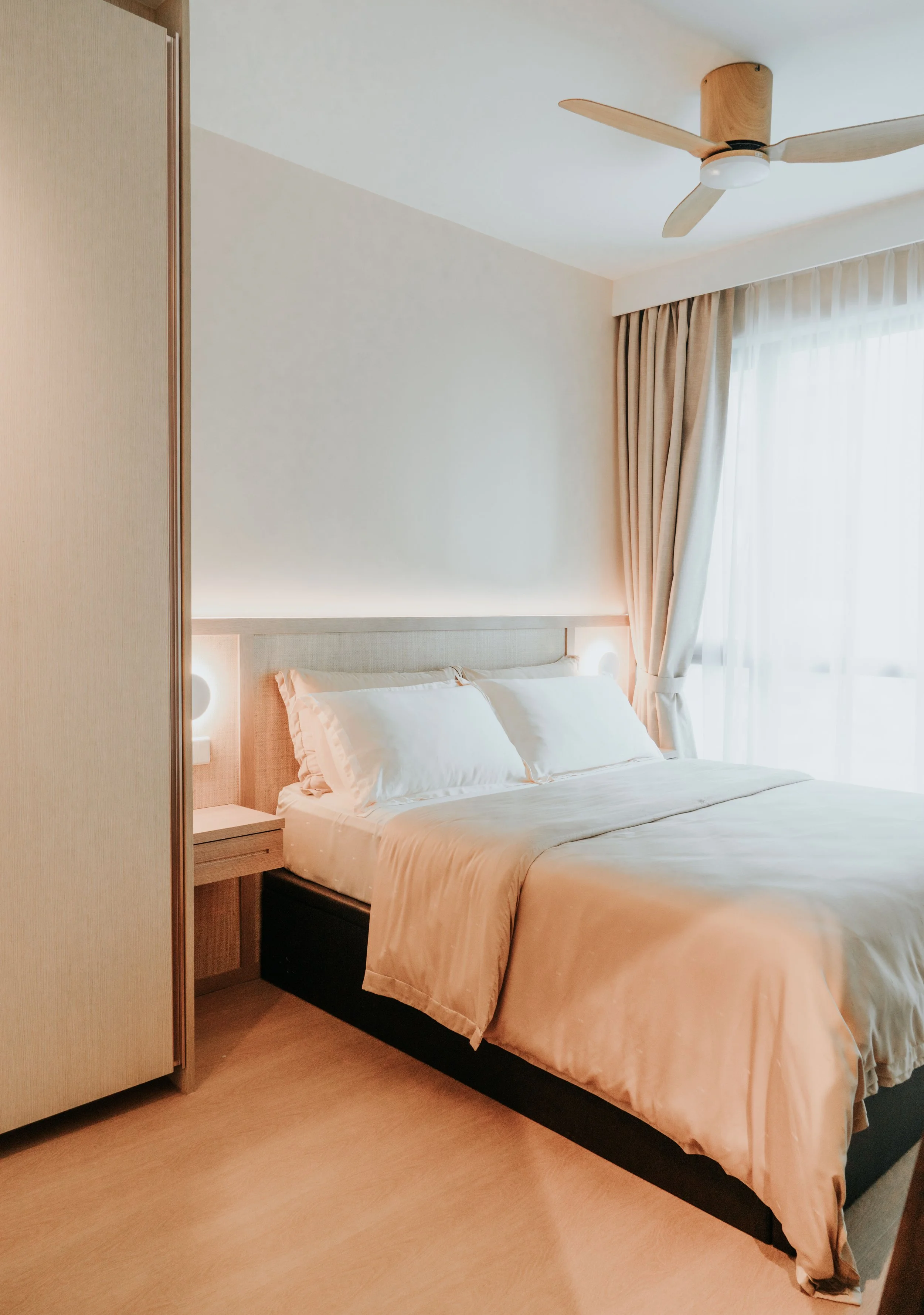 A neatly made bed with white bedding and pillows in a hotel room, featuring a bedside table, wall lights, curtains, and a ceiling fan.