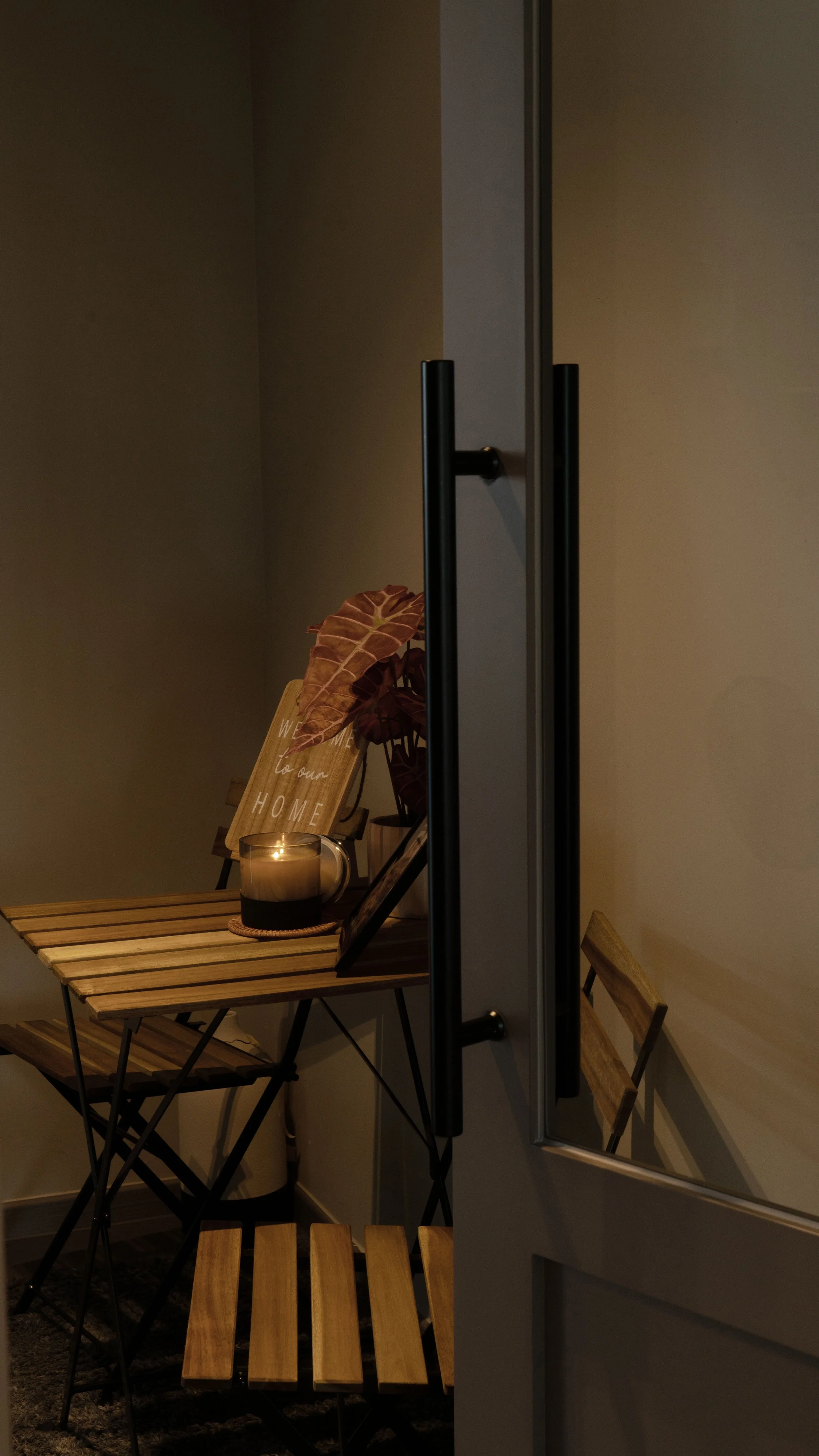 A cozy corner with a wooden table and chair, a lit candle in a decorative holder, a framed photo, and a welcoming sign with leaves, with minimal lighting creating a warm atmosphere.