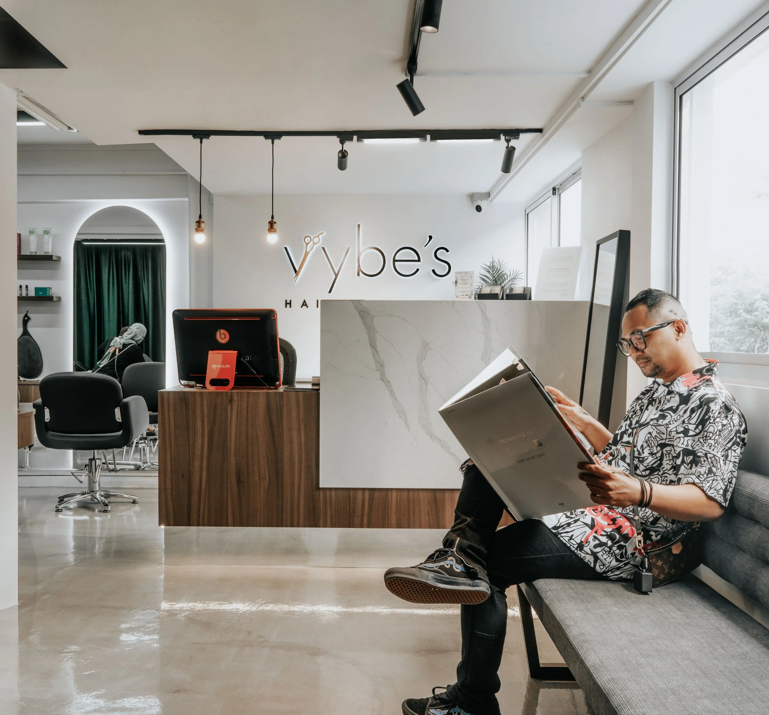 Man sitting on a gray bench reading a large book inside a modern hair salon with a reception desk and the sign 'Vybe's Hair' behind him.