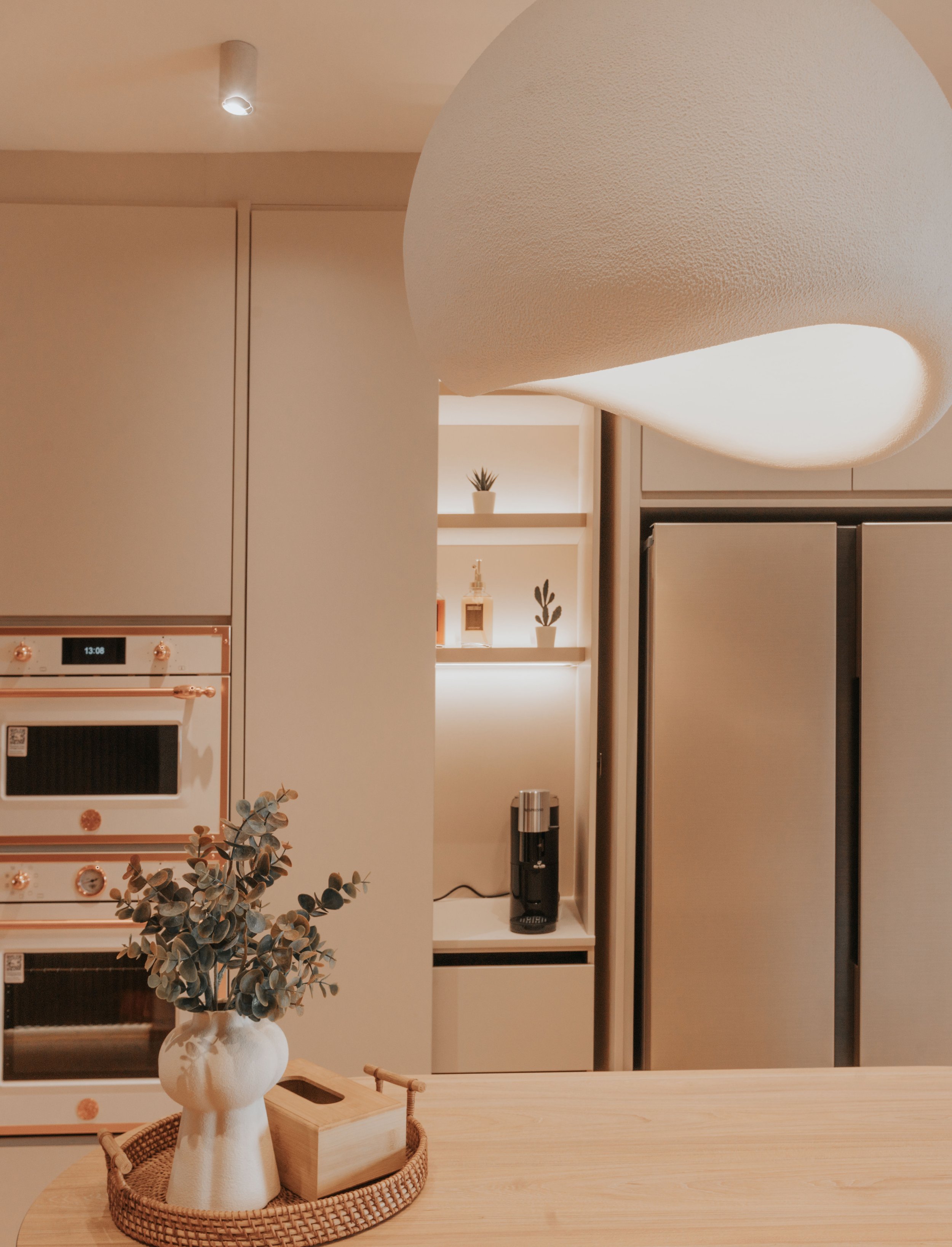 Modern kitchen with wooden countertop, decorative vase with eucalyptus, small tray, built-in oven, and in the background a coffee machine, refrigerator, and shelves with small potted plants and bottles, illuminated with warm lighting.