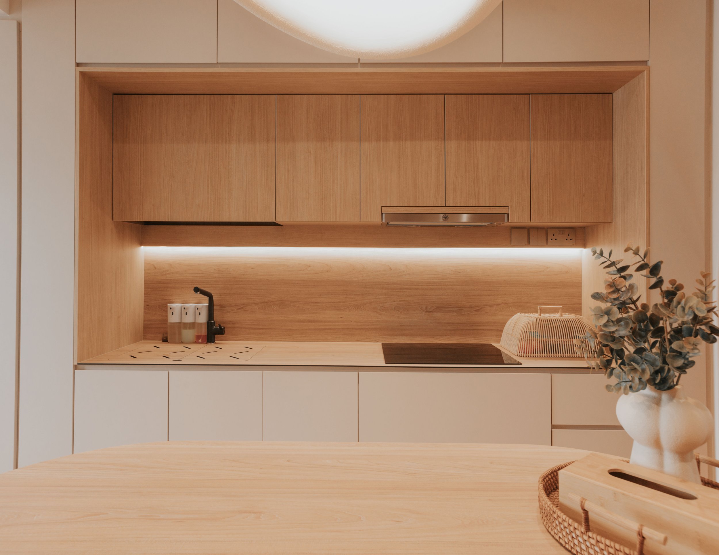 Minimalist kitchen with wooden upper cabinets, white lower cabinets, black faucet, and a wooden table with a vase of eucalyptus.