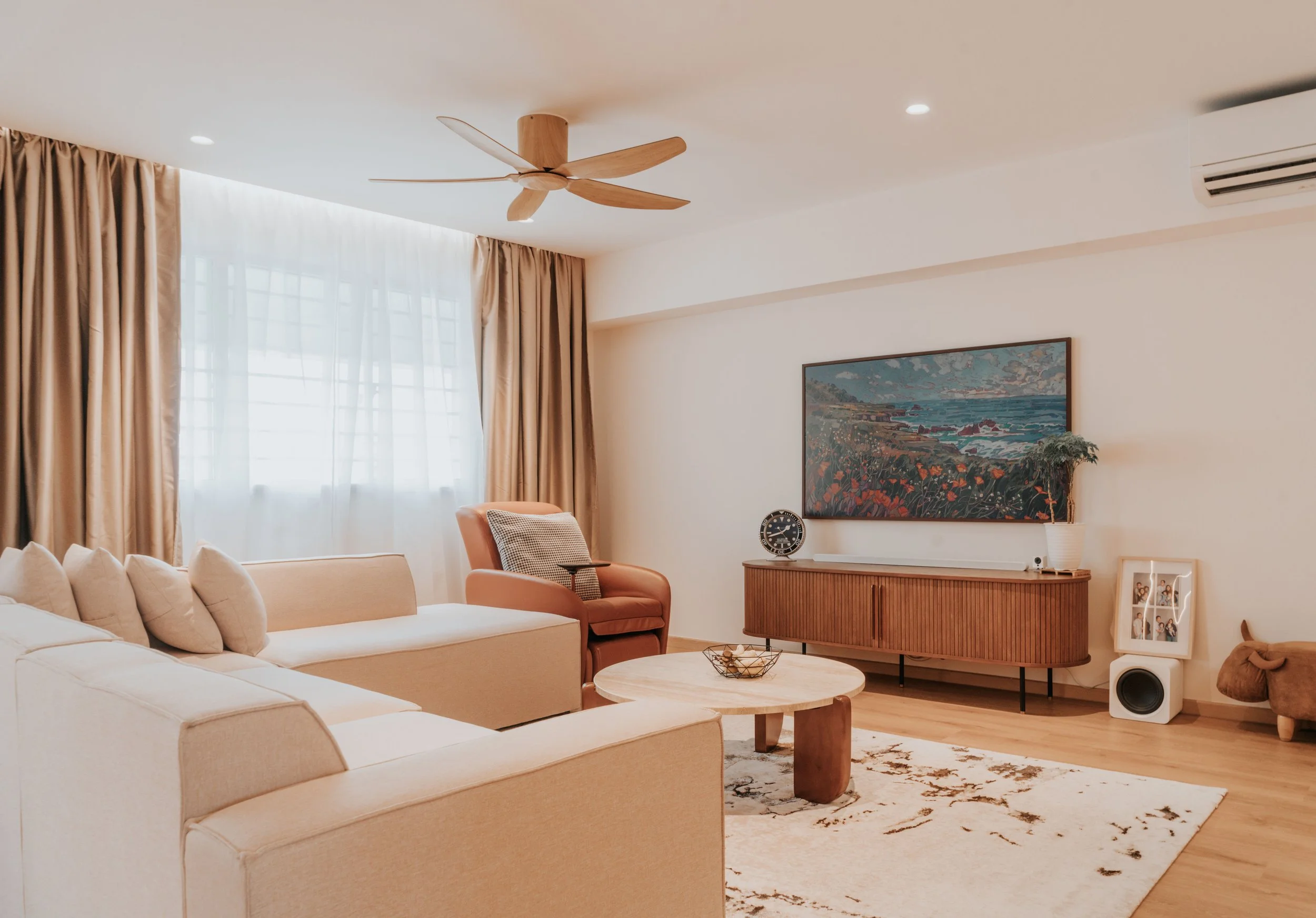A living room with beige sofas, a pink armchair, a wooden coffee table, and a TV console with wall art above. There are big windows with sheer white curtains and beige drapes, and a ceiling fan.