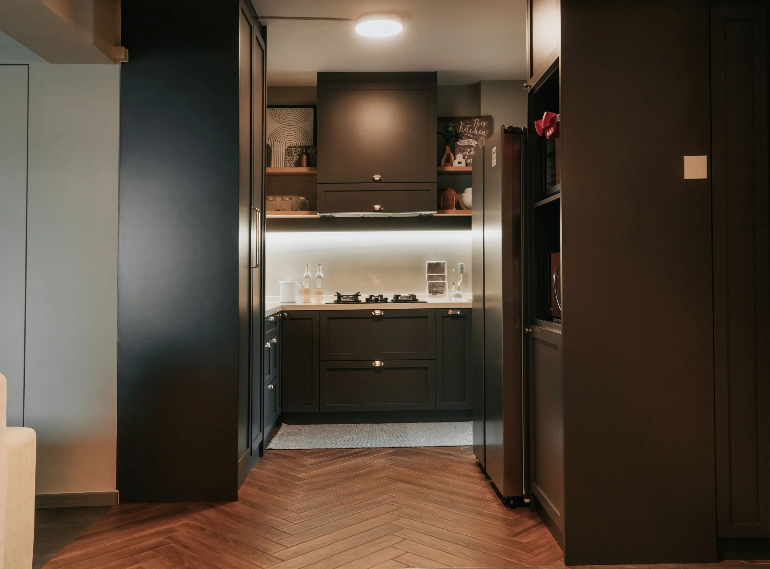 Modern kitchen with dark cabinets, a small countertop with bottles and a stove, under-cabinet lighting, and wooden herringbone-patterned flooring.