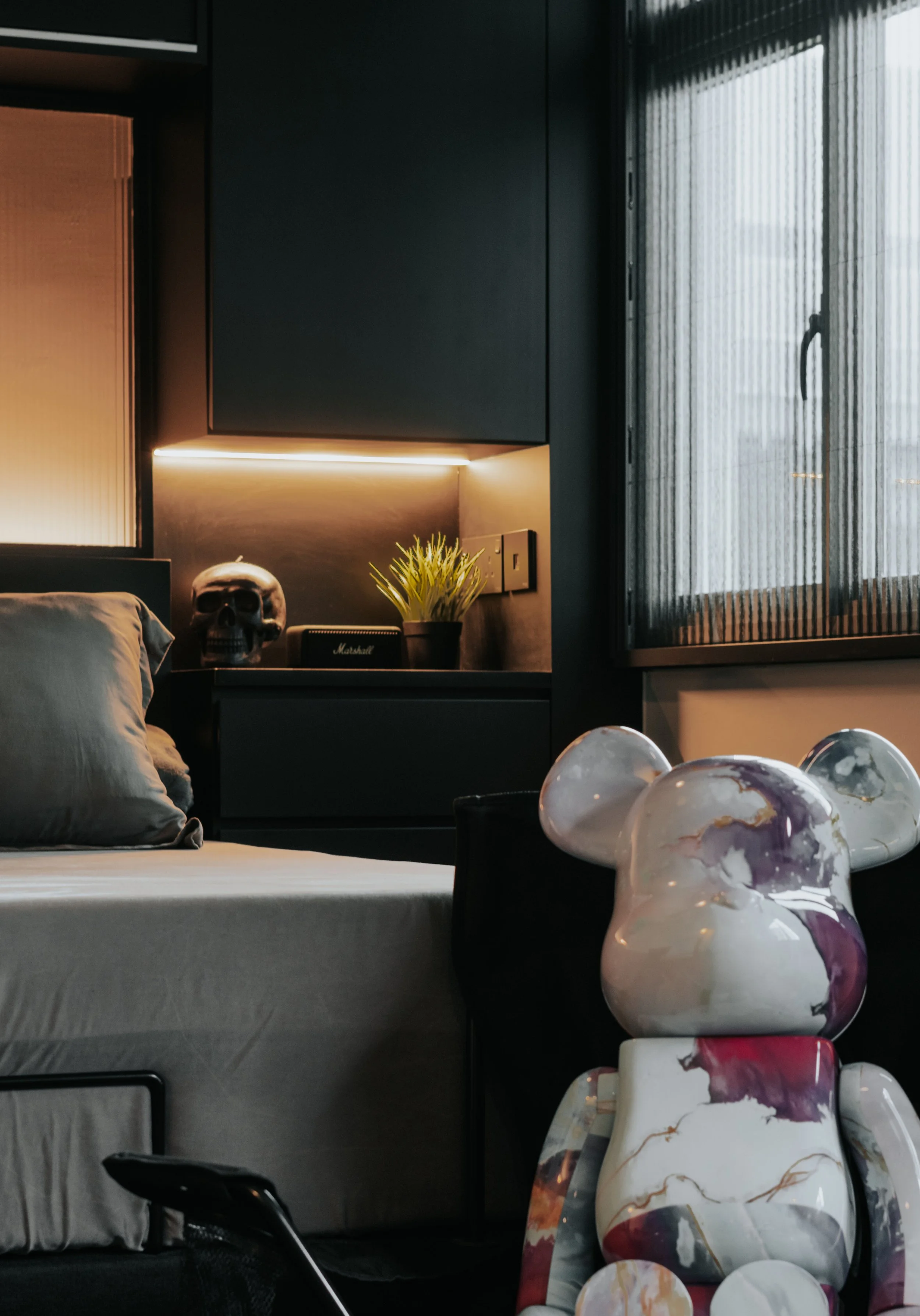 Part of a modern bedroom with a bed, decorative skull, small potted plant, and a colorful teddy bear sculpture near a window with black blinds.