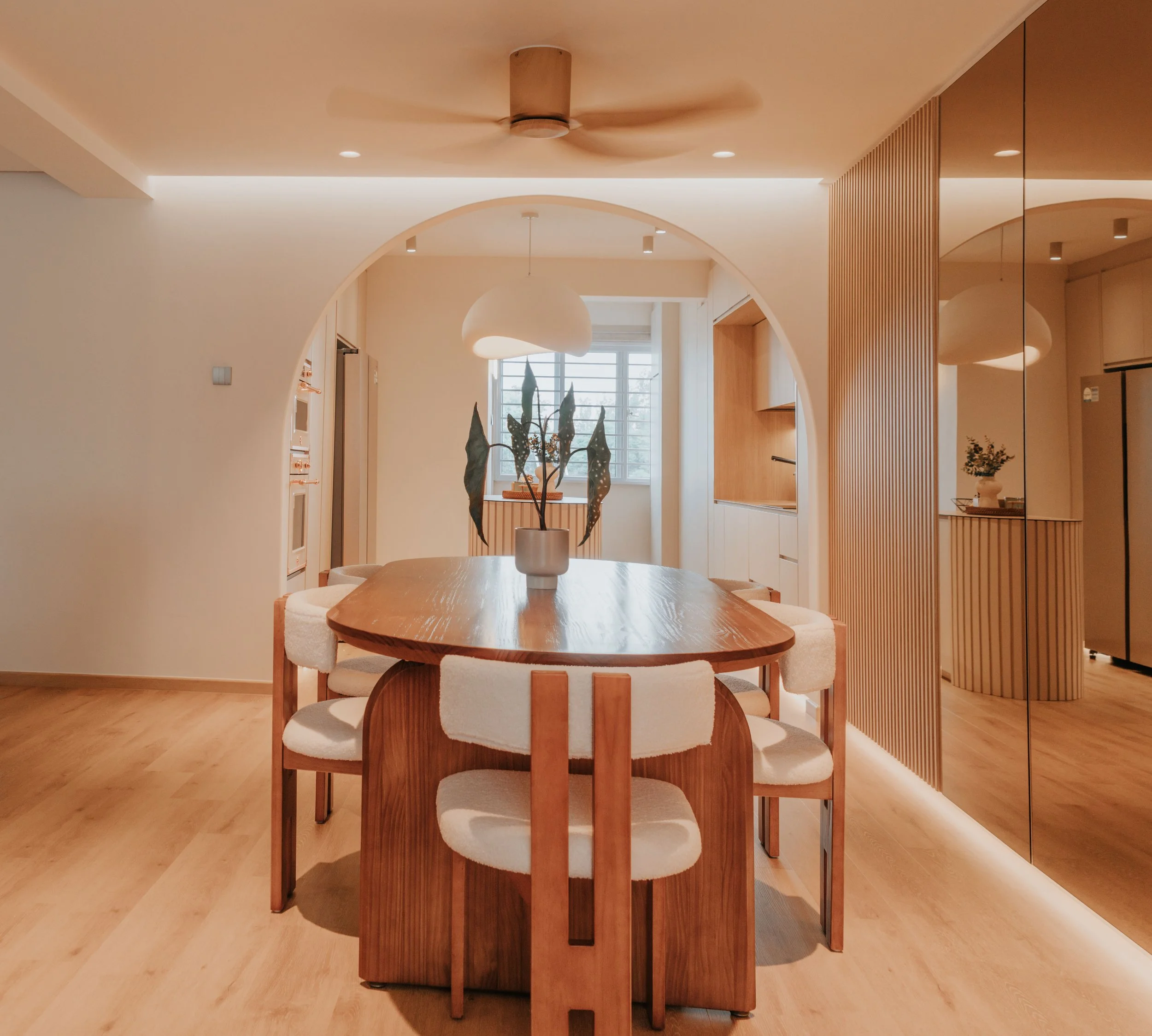 Modern dining area with wooden table, six upholstered chairs, potted plant centerpiece, wood and mirrored accents, and natural light from window.