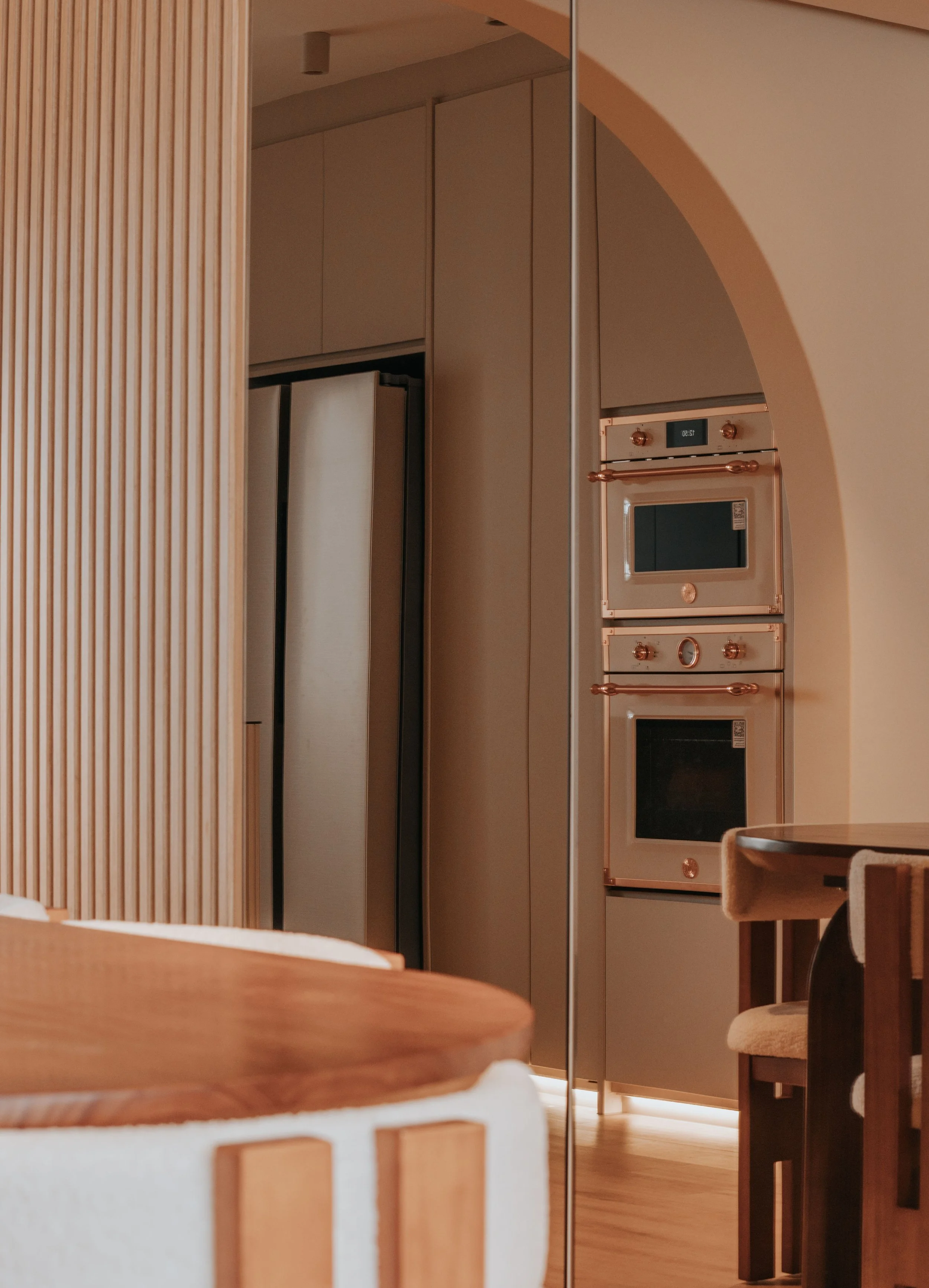 View of a modern kitchen with built-in oven and microwave in a metallic finish, via an arched doorway, with wooden tables and chairs visible in the foreground.