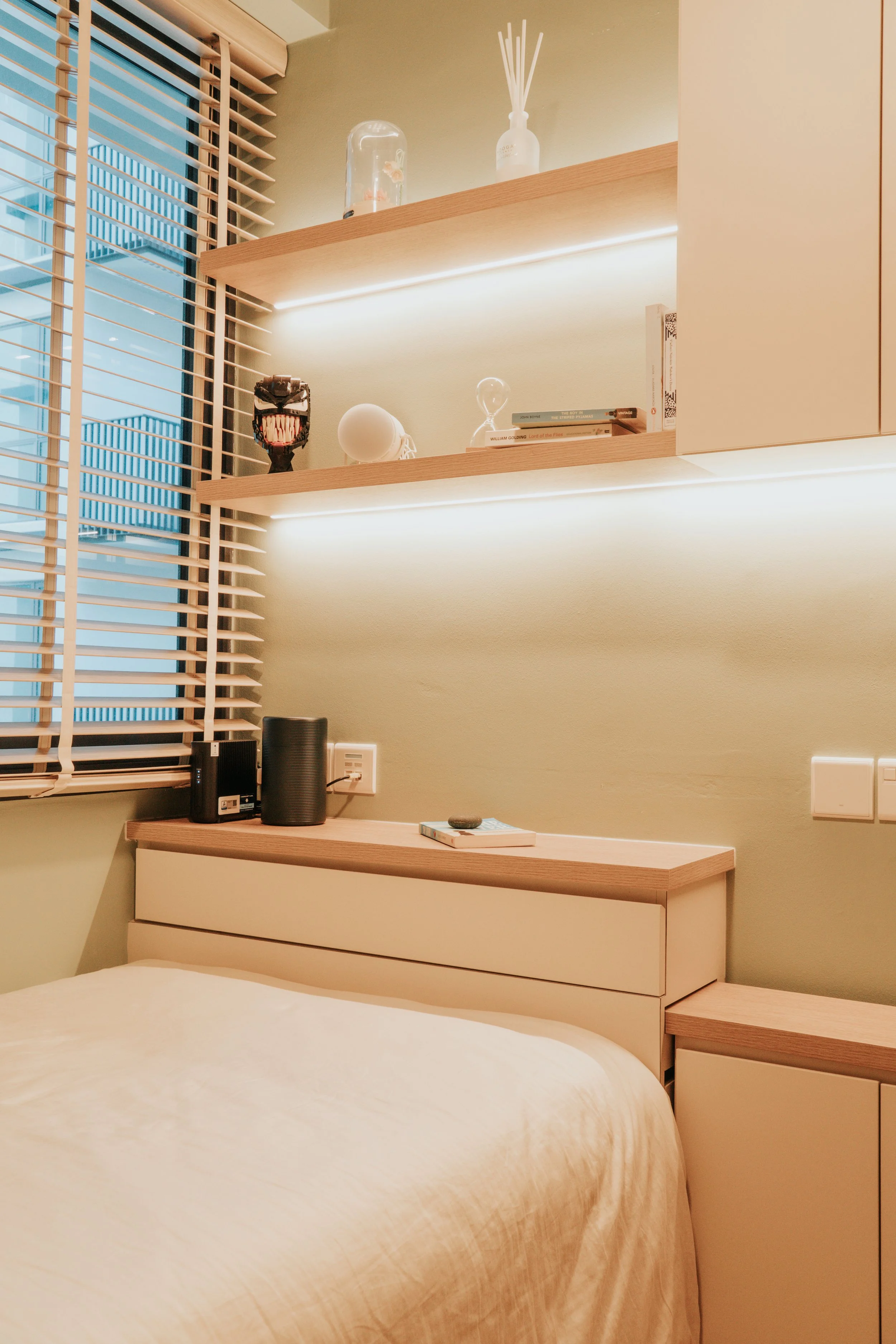 A cozy bedroom corner with a bed, a wooden headboard, and a shelf with decorative items, near a window with wooden blinds.