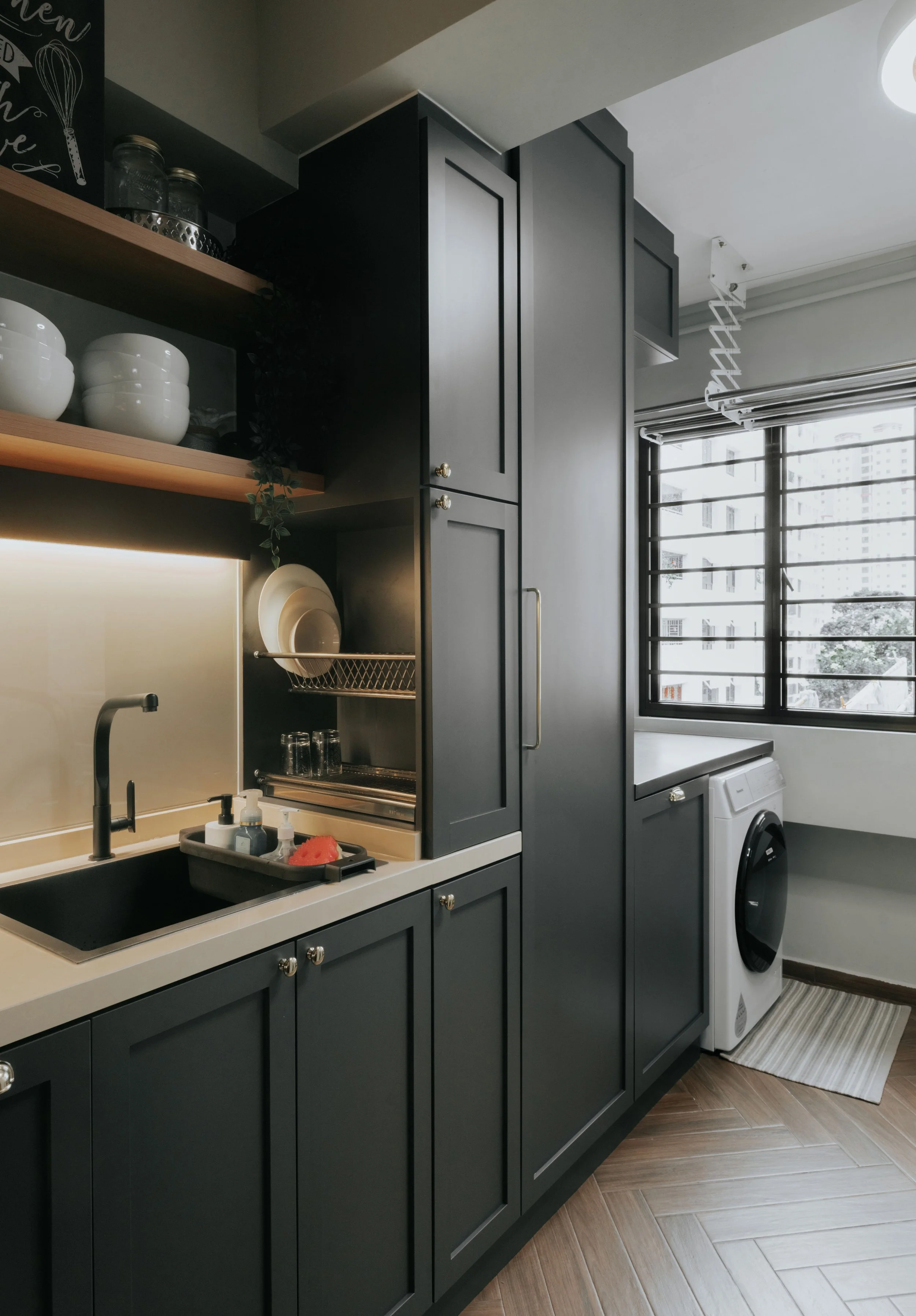 Modern kitchen with black cabinets, open shelving with white dishes, a black sink, a dish drying rack, and a washing machine near large windows with black frames.