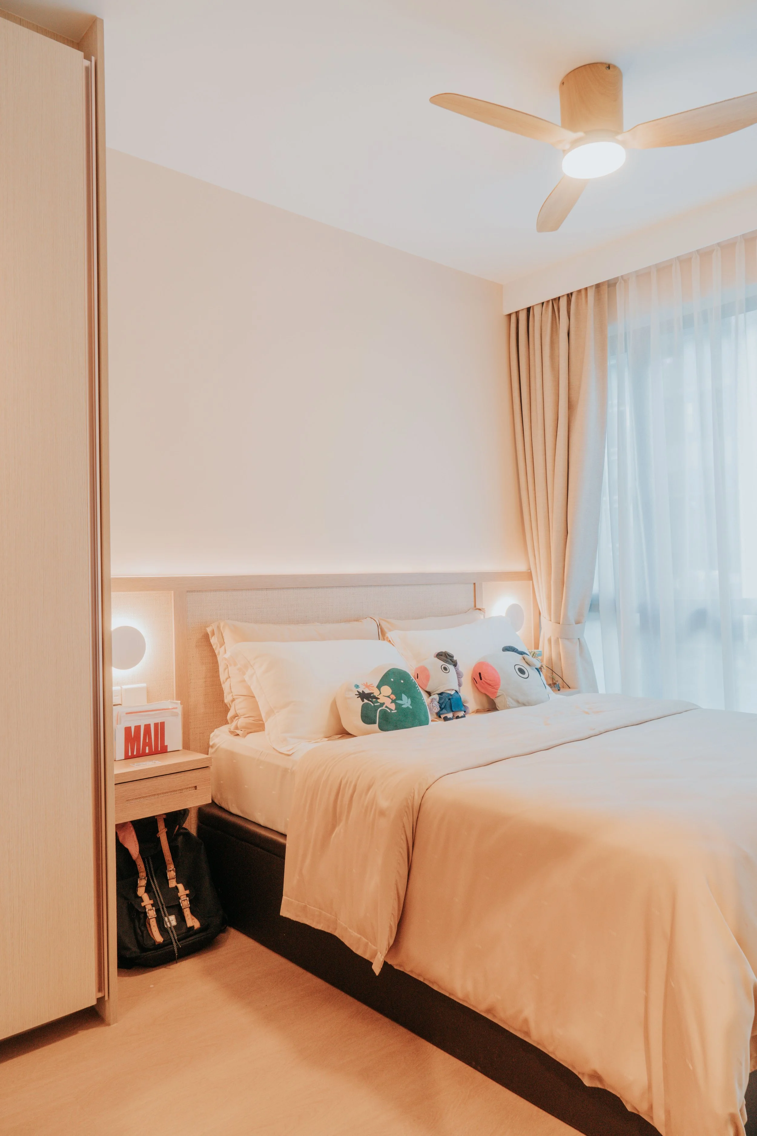 A neatly made bed with cream-colored bedding and decorative pillows featuring cartoon characters, next to a wooden nightstand with a mailbox-shaped decoration labeled 'MAIL' and a backpack underneath, in a room with white walls, beige curtains, and a